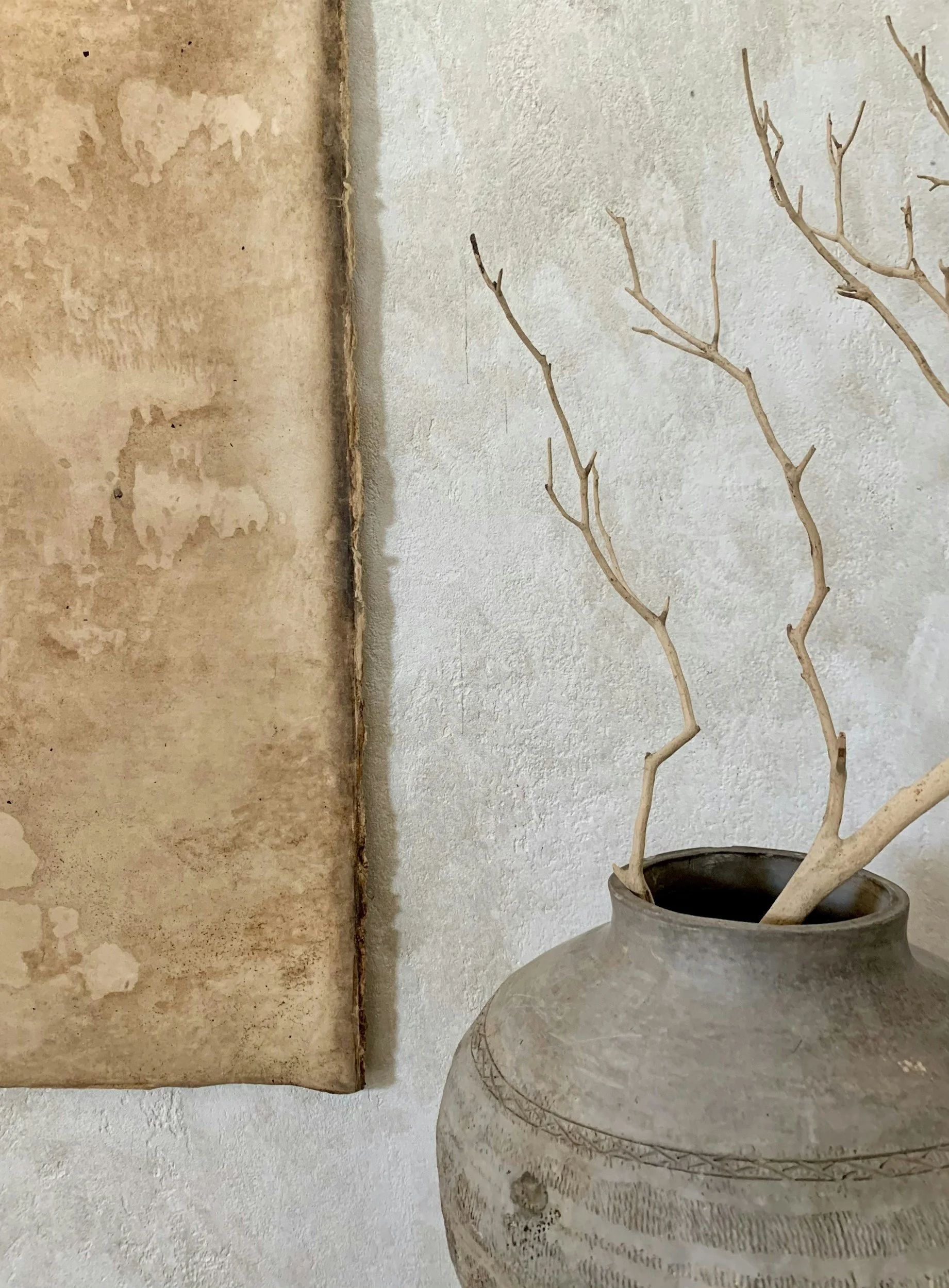 A large decorative pottery vessel with carved patterns, holding a dried plant with bare branches, positioned in a corner next to a beige textured wall and a weathered, painted wall.