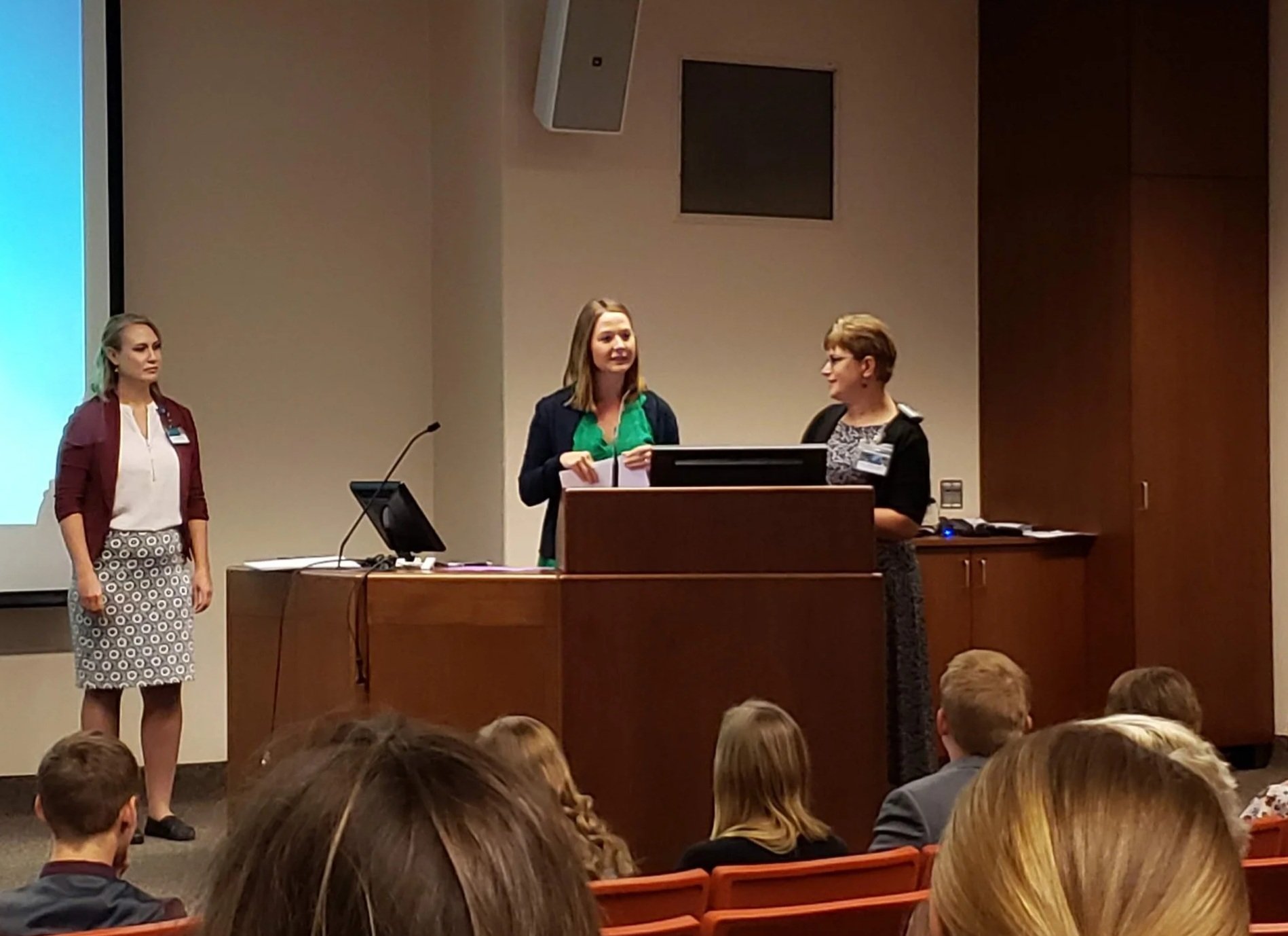 Three women standing at a podium in front of an audience during a presentation or conference in a conference room. One woman is speaking, while the other two are listening.