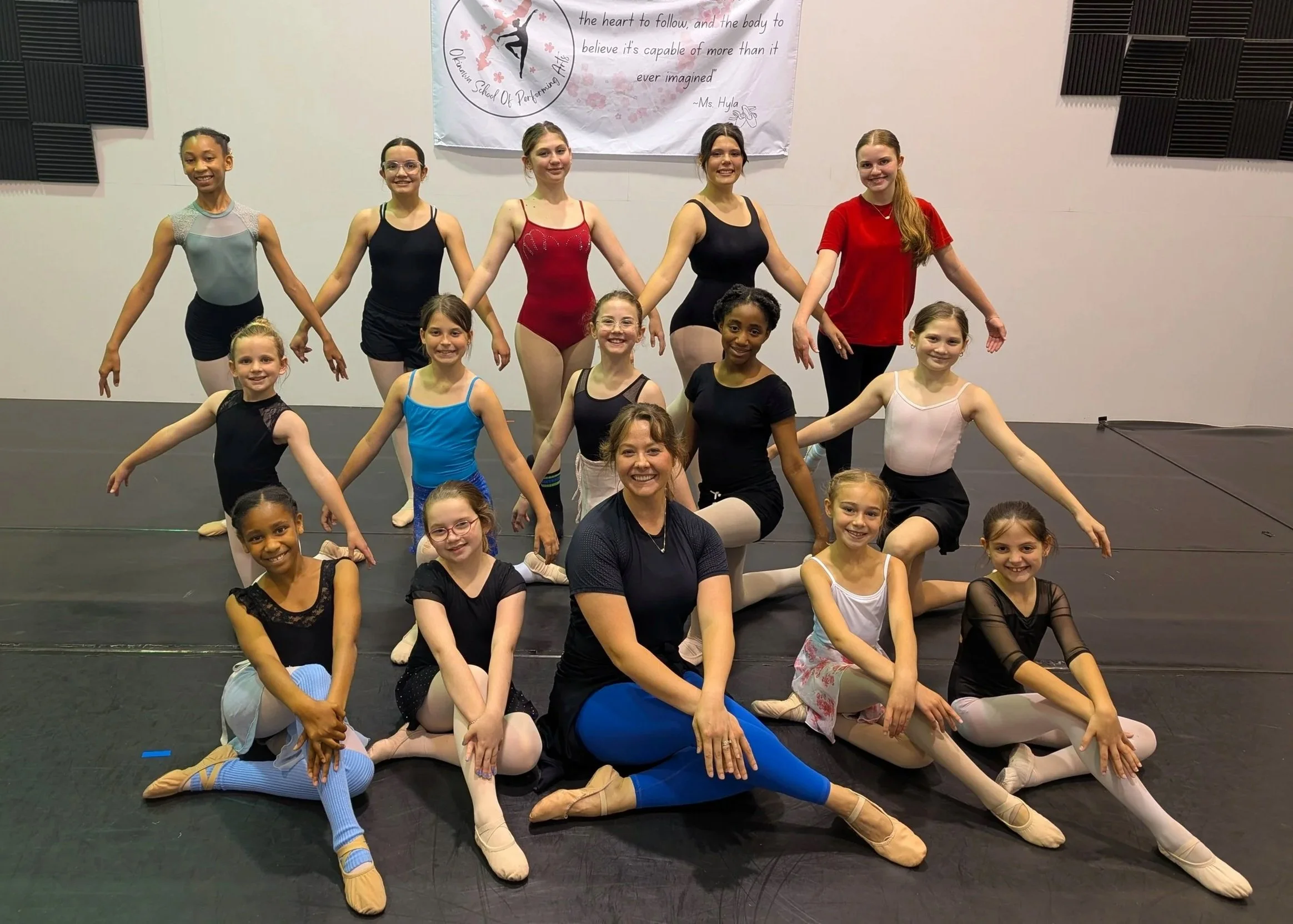 Group of young girls in ballet costumes and two adult women posing together in a dance studio, smiling at the camera. The studio has a banner on the wall with a dance-related quote and black soundproofing panels.