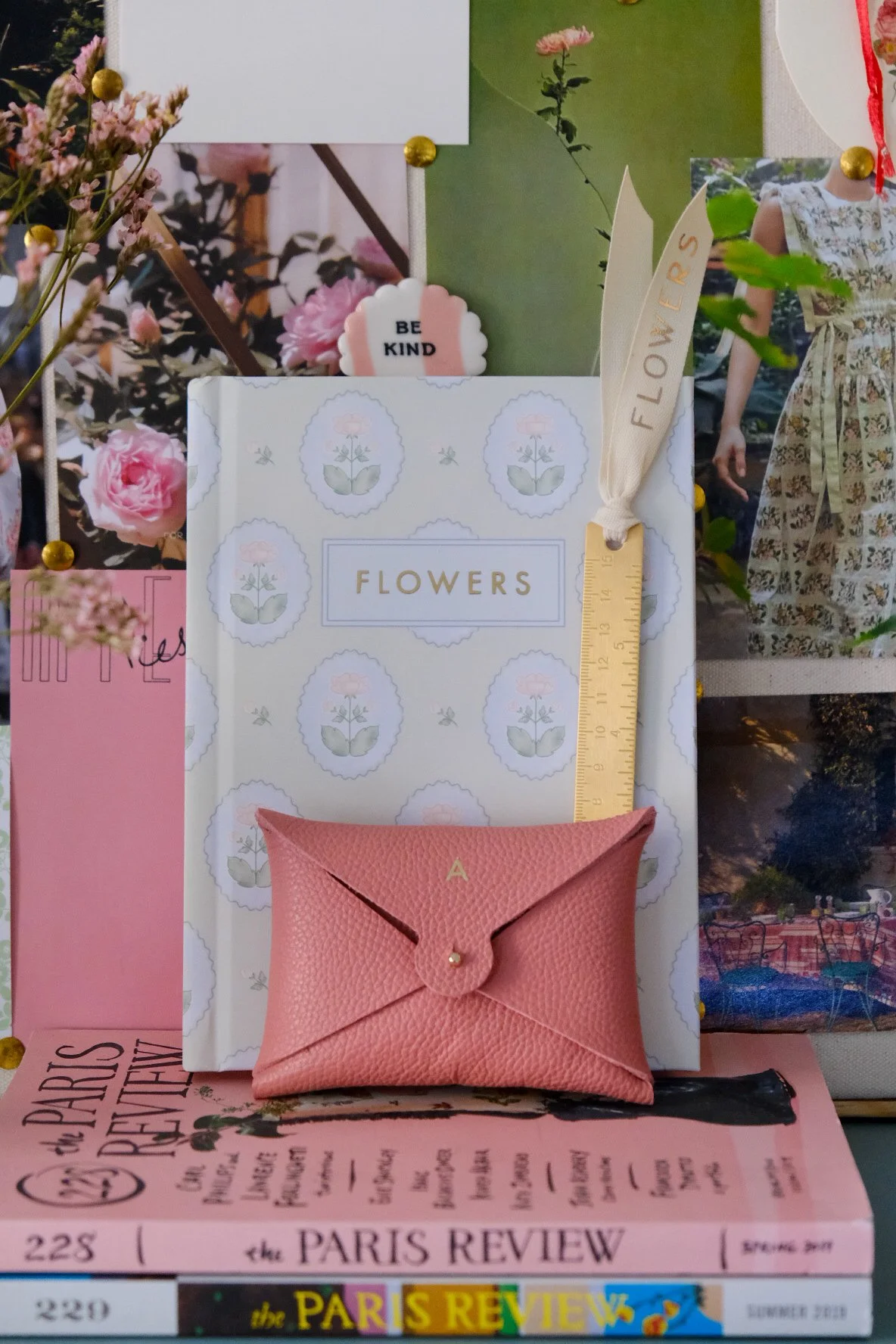 A display of pink and white flowers, a box labeled "FLOWERS," a small pink leather pouch with a gold letter "A" on it, a ruler marked in inches, and various photos and notes on a bulletin board.