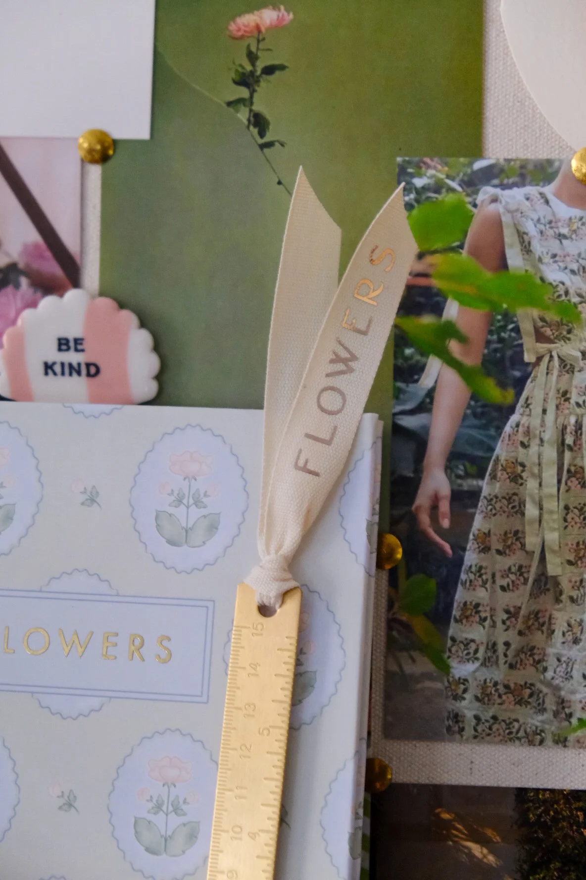 A close-up of a collage or scrapbook with a yellow ribbon labeled 'FLOWERS' attached to a white card that has a pattern of light blue floral outlines and the text 'FLOWERS' in gold. The collage features a photograph of a young girl in a floral dress and greenery, a small pink flower, a pink and white 'BE KIND' sign, and a small picture of a pink flower.