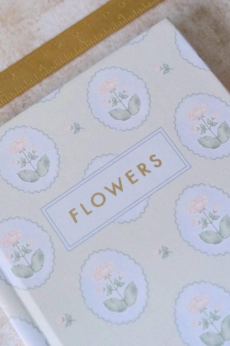 A book titled 'Flowers' with a light-colored cover decorated with pastel pink roses and green leaves, beside a yellow measuring tape.