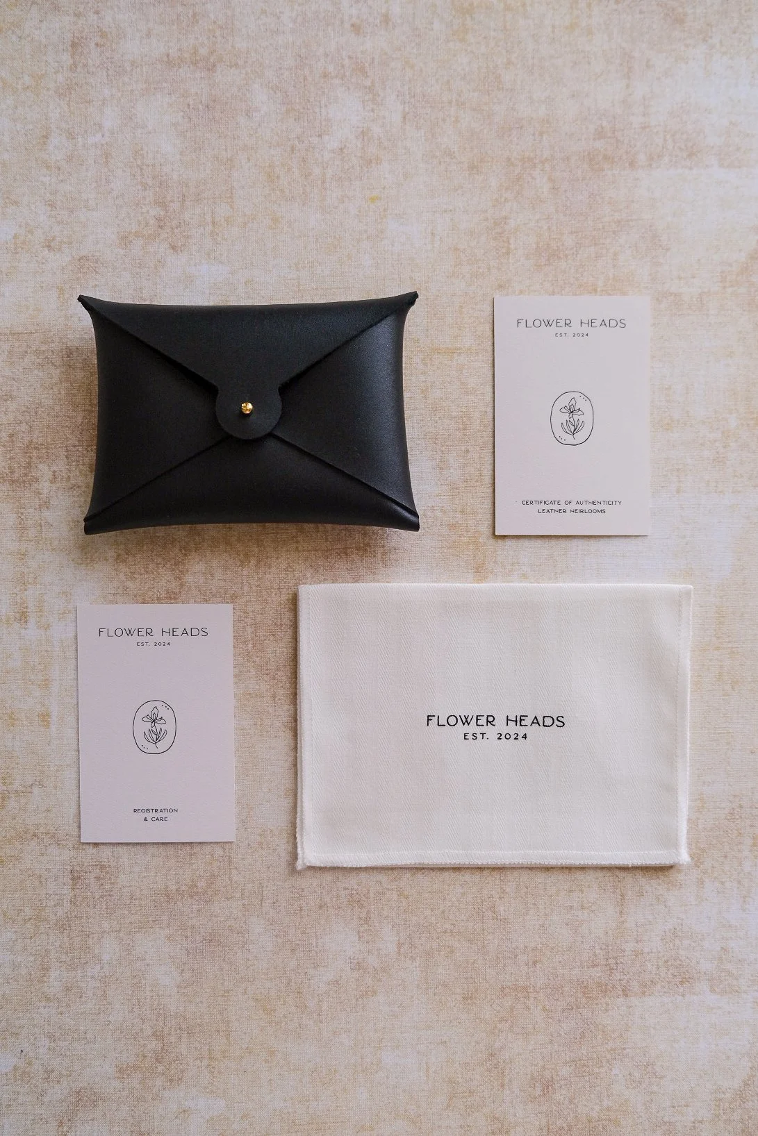 Black leather wallet with gold button, white paper cards with black text and logo, and a white cloth with black text, all branded 'Flower Heads' arranged on a beige textured surface.