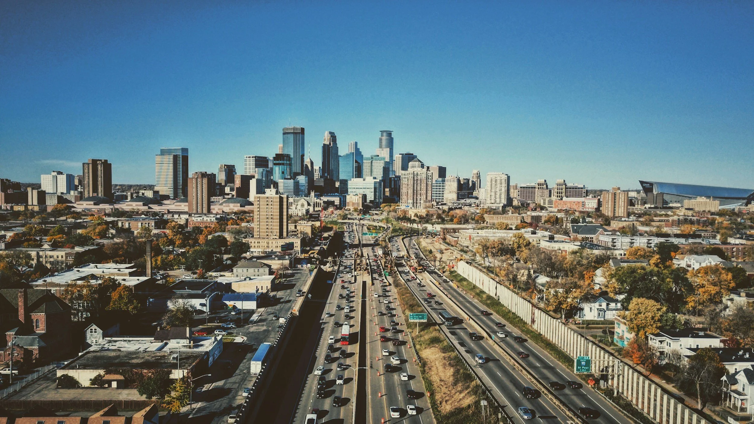 A city skyline with tall buildings and skyscrapers under a clear blue sky, with a busy highway filled with cars and trucks in the foreground.