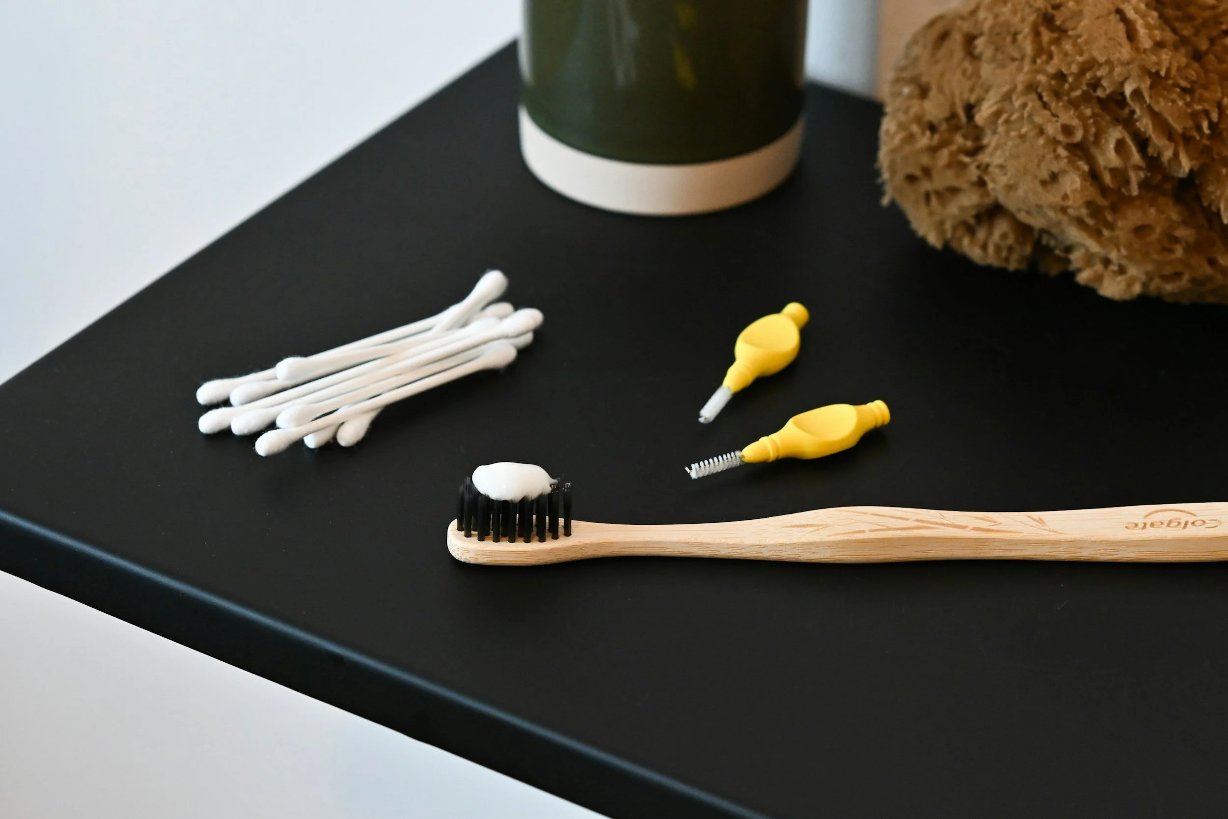 A black table with cotton swabs, a yellow ear cleaning tool, a black brush with white bristles, a green and white mug, and a brown textured sponge.