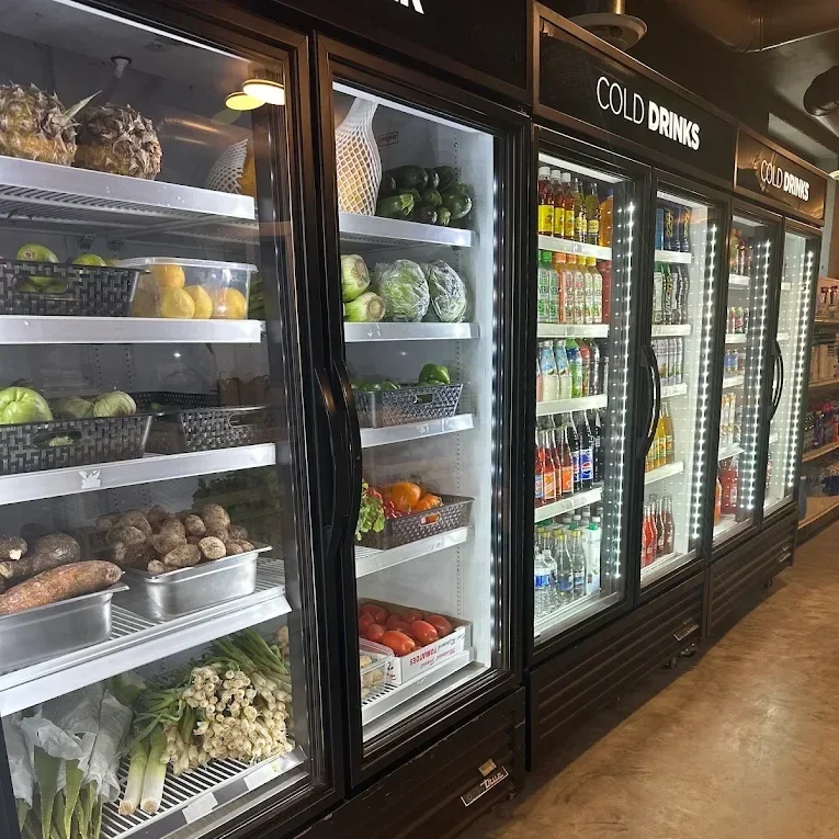 Refrigerator with fresh fruits and vegetables on the left, including pineapples, green peppers, lettuce, oranges, tomatoes, and green onions. The middle and right sections contain bottled drinks and beverages.
