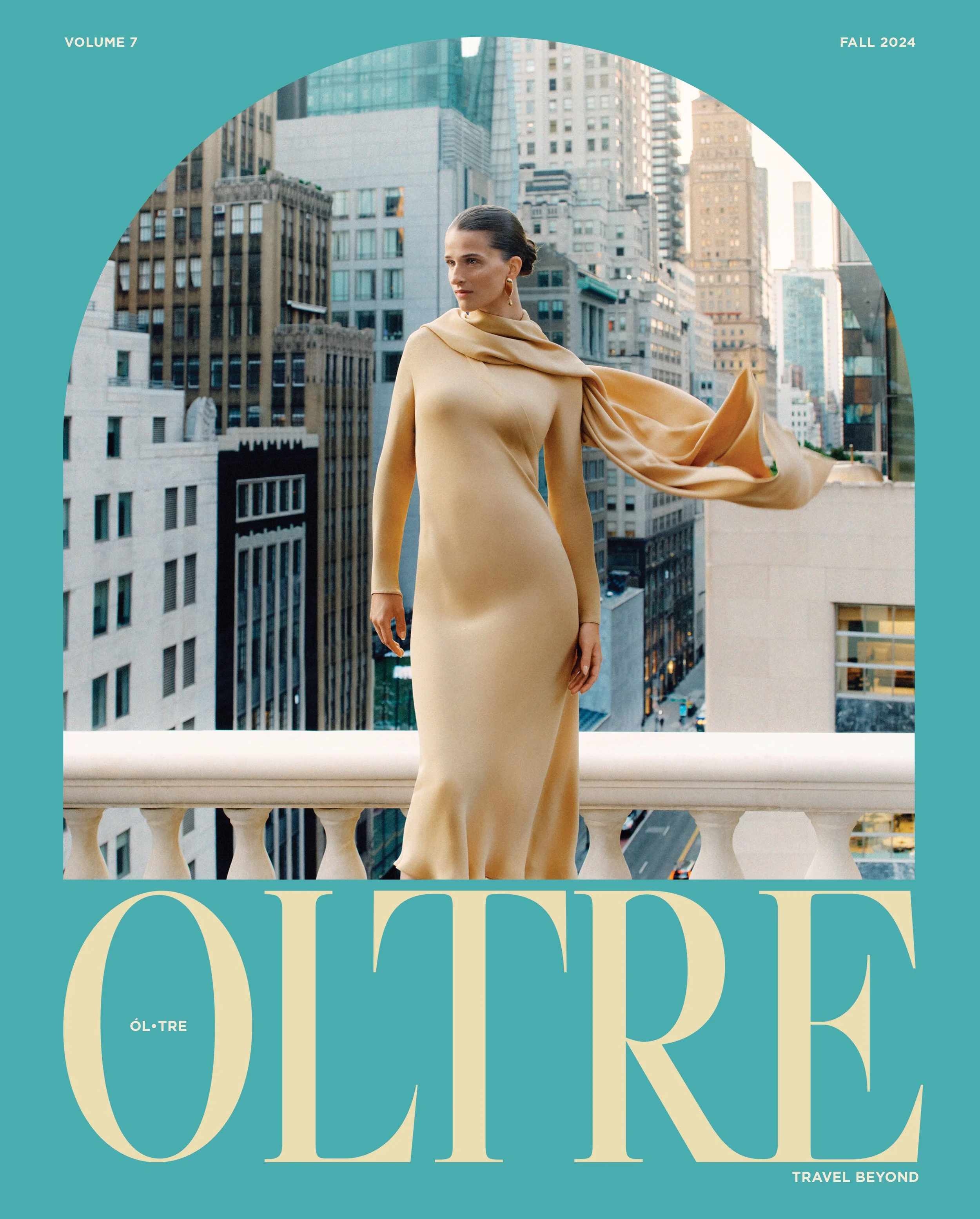 A woman in a beige dress with a matching scarf draped over her shoulder standing on a balcony with a cityscape background. The image is part of a magazine cover for the fall 2024 issue of OLTRE, Volume 7. The cover features large stylized text readin
