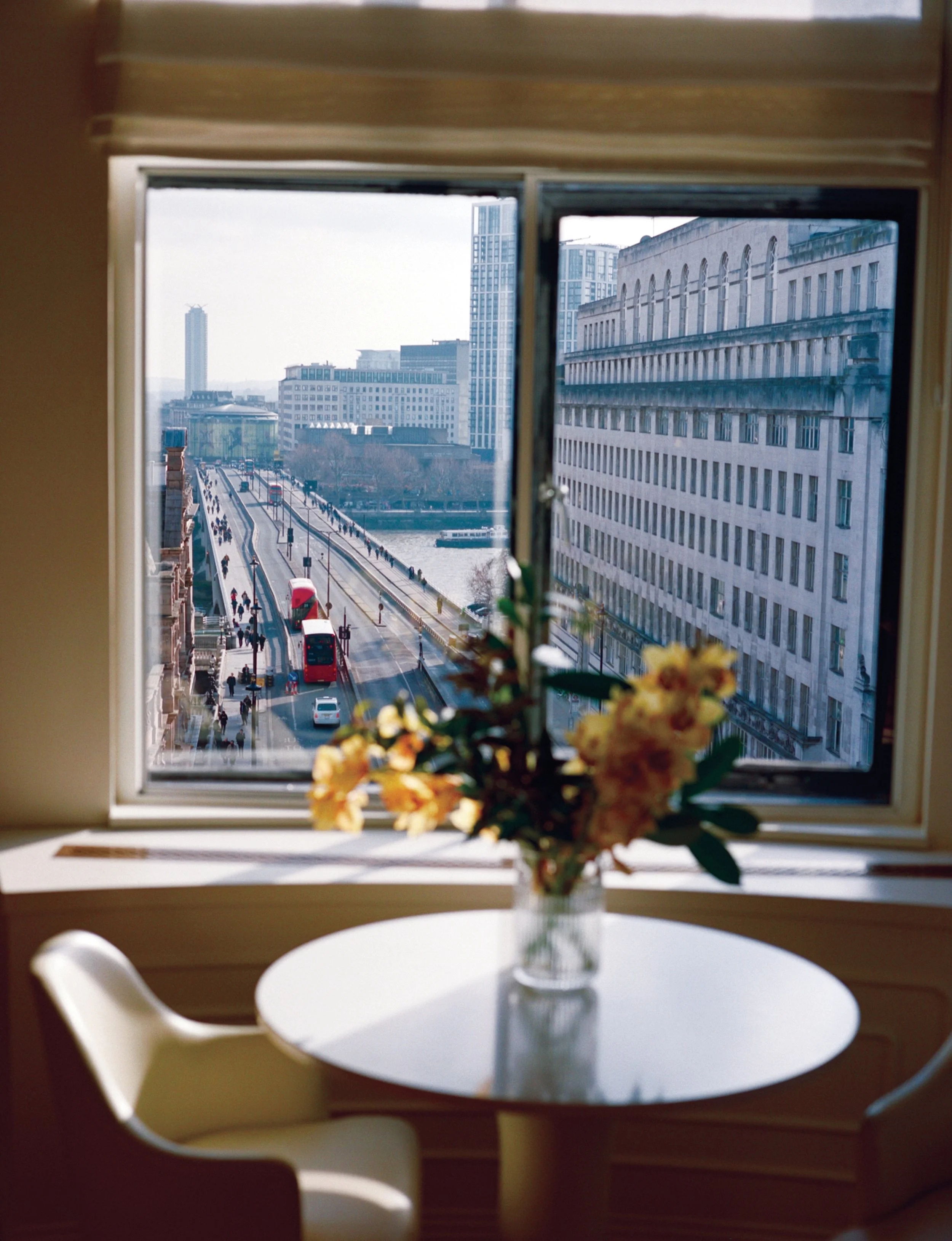 A vase with flowers on a white round table, viewed through a window showing a city street with tall buildings and buses.