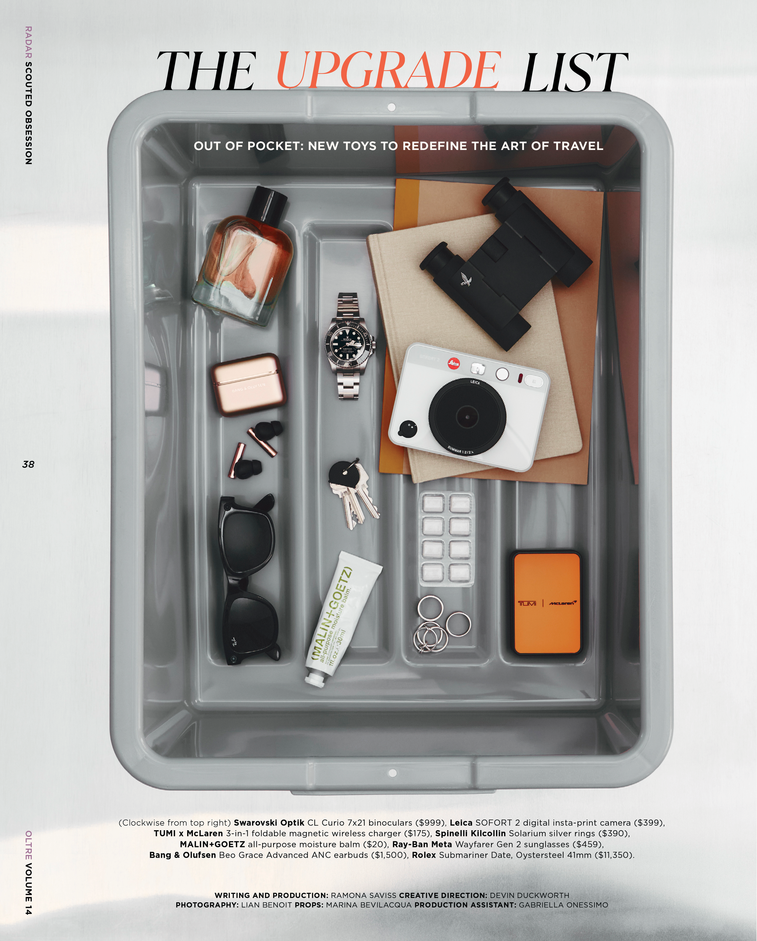 A gray tray containing various items: a brown bottle of perfume, an Apple Watch, a Leica camera, a beige notebook, binoculars, a wireless charger, a tube of balm, a pair of sunglasses, a few rings, and a set of keys. The image is part of a magazine s