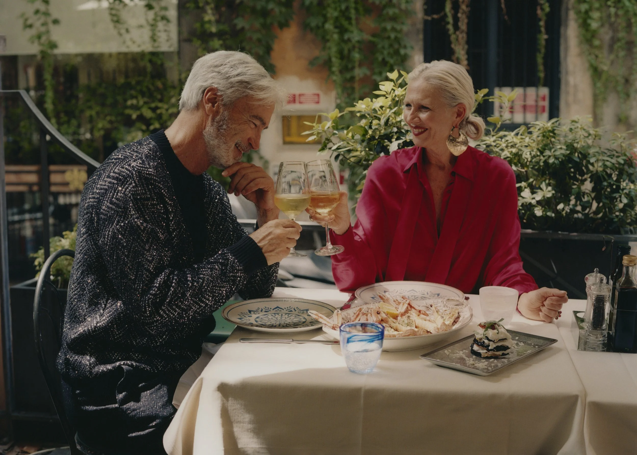 An elderly man and woman sitting at a table outdoors, sharing a toast with wine glasses and smiling. The table has a plate of lobster and a small cake. Background includes greenery and outdoor seating.