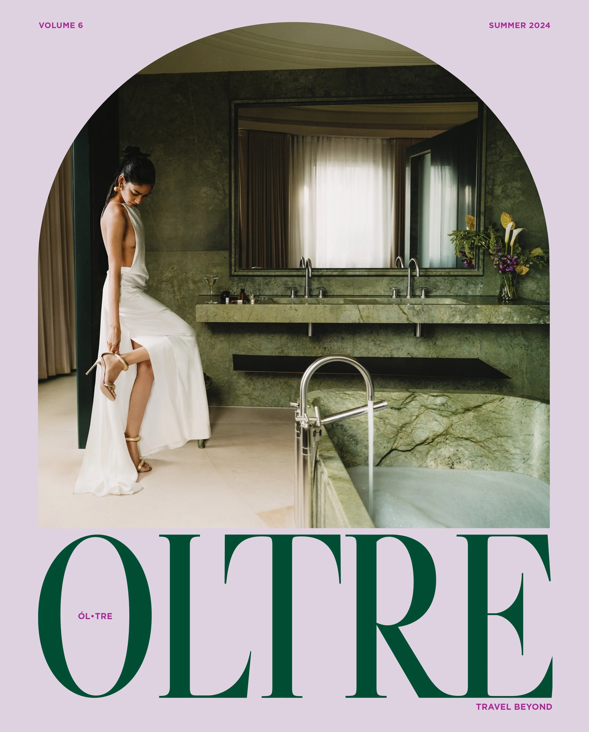 A woman in a white dress and high heels stands in front of a large mirror in a luxurious bathroom with green marble surfaces, flowers, and two sinks.