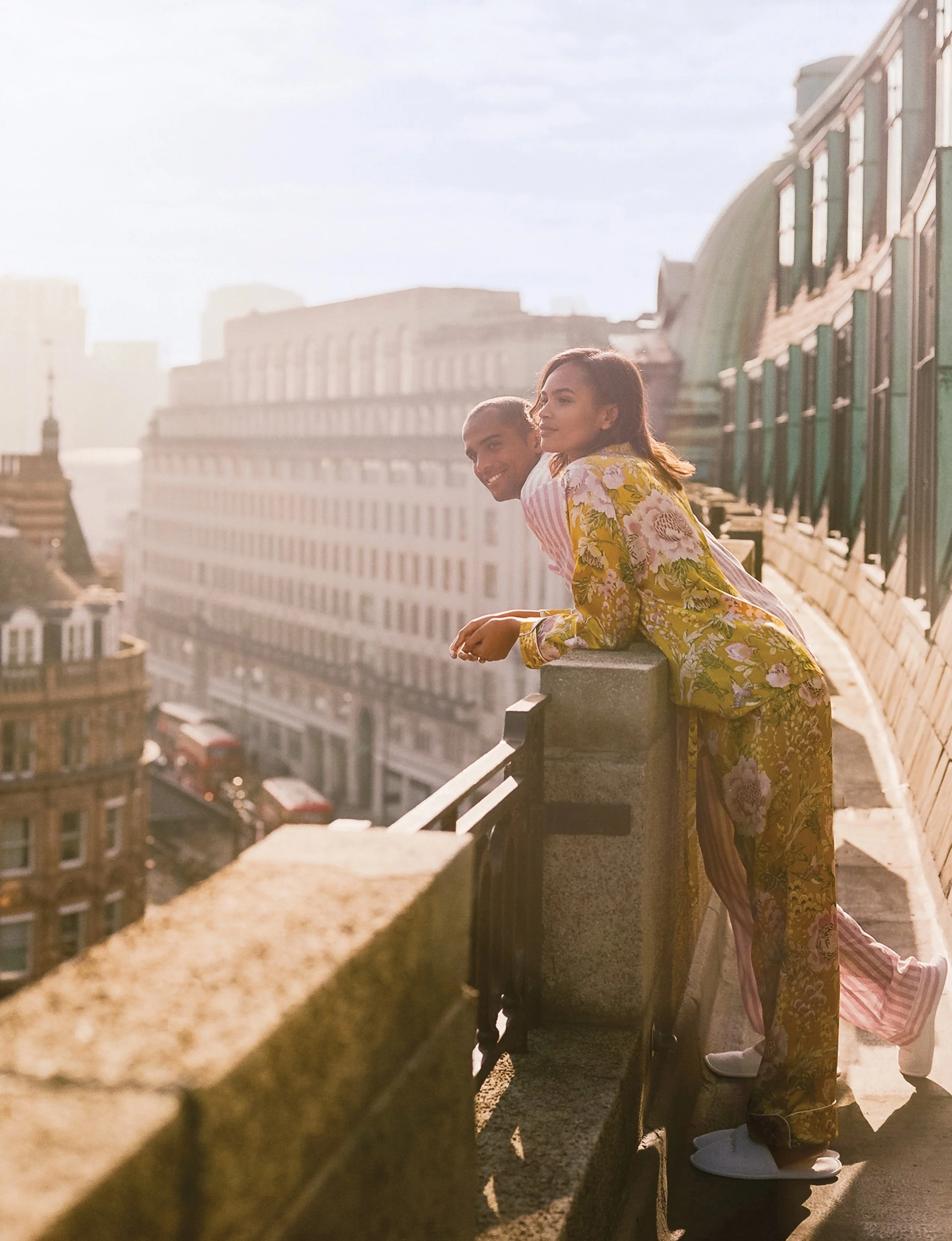 Two women leaning on a rooftop railing overlooking a cityscape in the late afternoon with sunlight casting a warm glow.