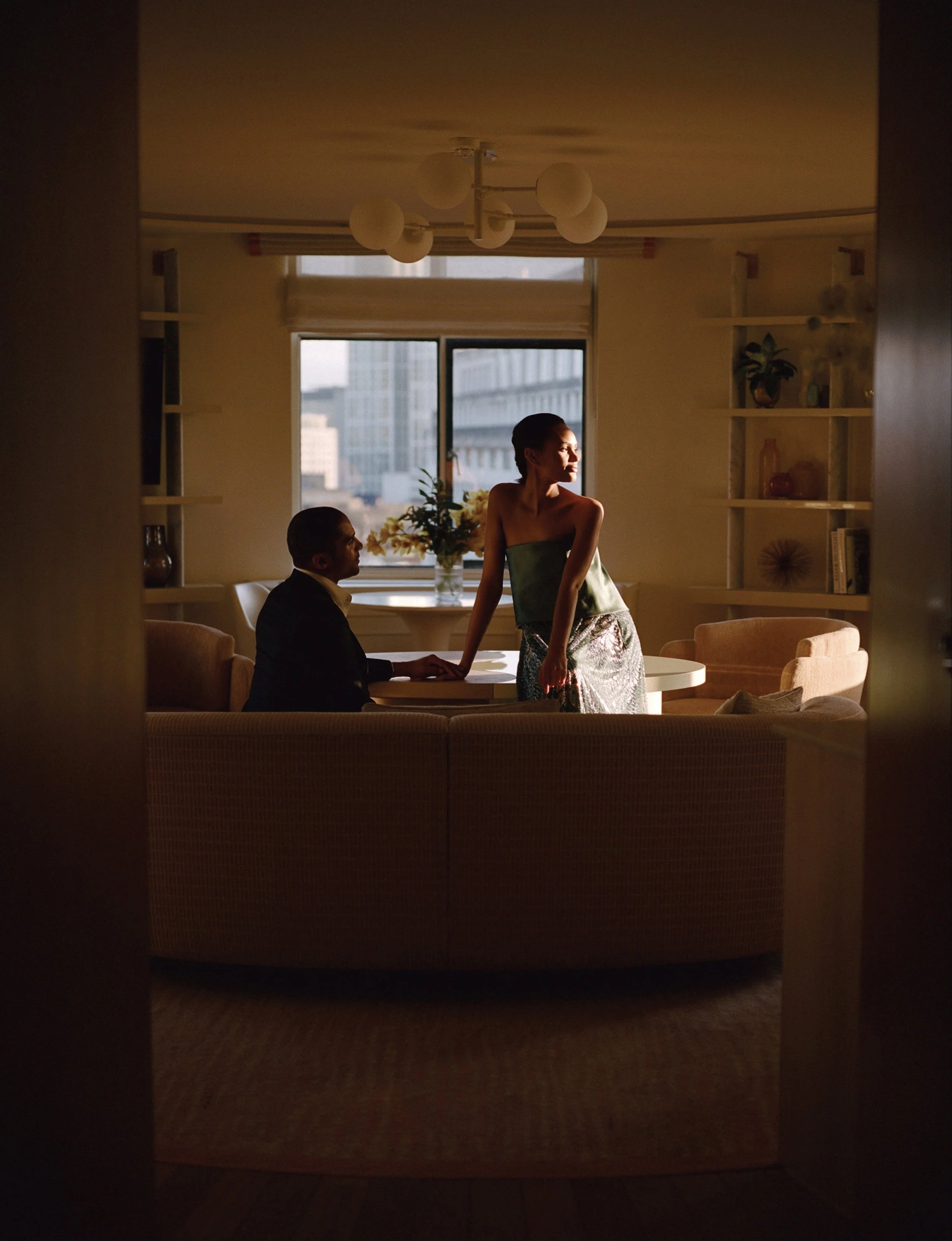 A woman and a man are sitting in a sunlit room with large windows. The woman is standing, facing to the right, while the man is seated, facing her. The room has shelves with plants and decorative items, and a vase of flowers on the windowsill.