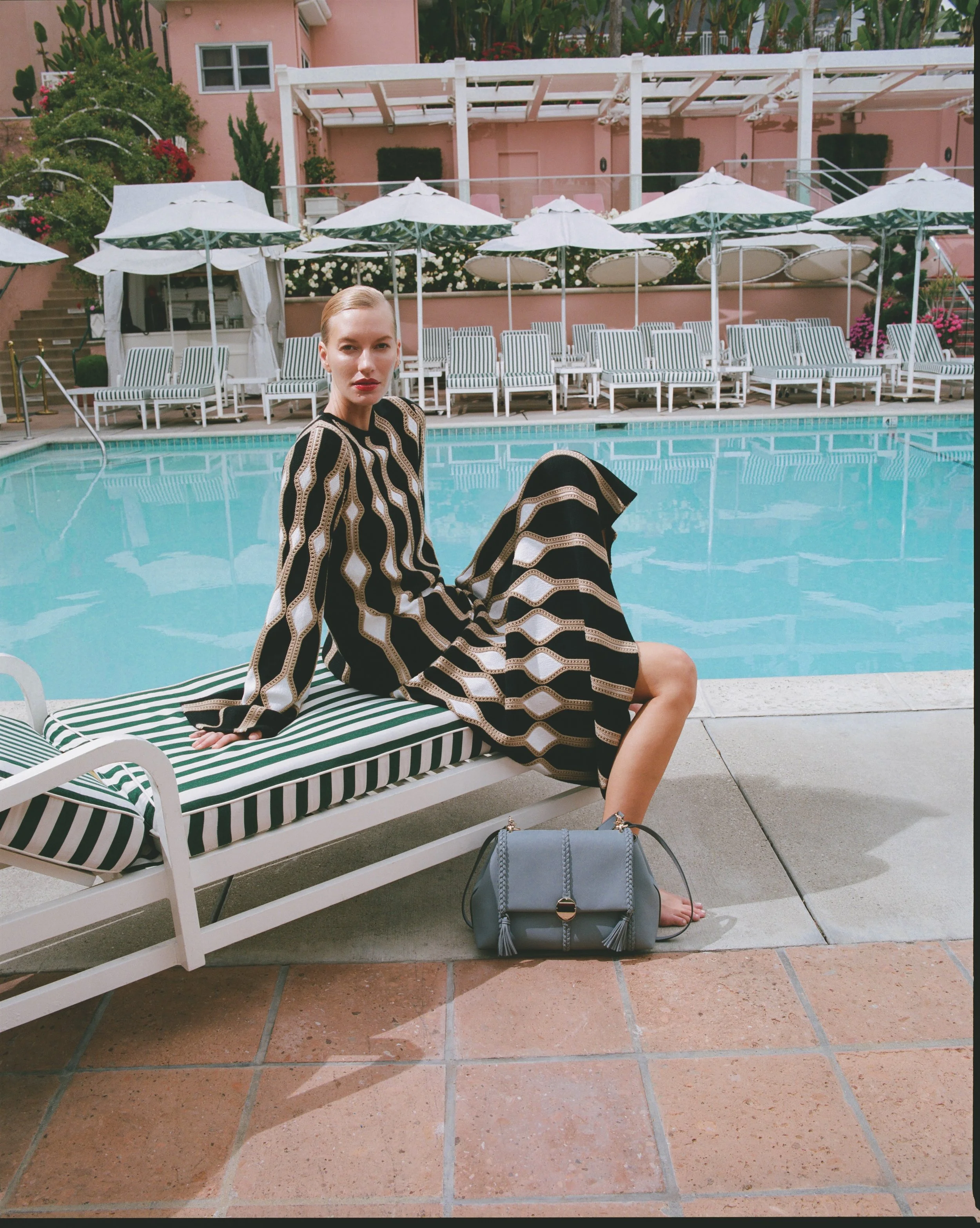 A woman sitting poolside on a striped lounge chair wearing a patterned dress, with a gray handbag on the ground, pool and white umbrellas behind her, pink building and greenery in background.