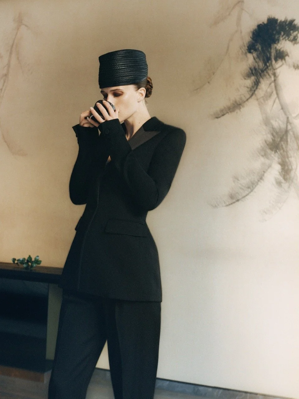 A woman dressed in a black suit with a matching hat is drinking from a black cup while standing in front of a beige wall with black abstract brush strokes.