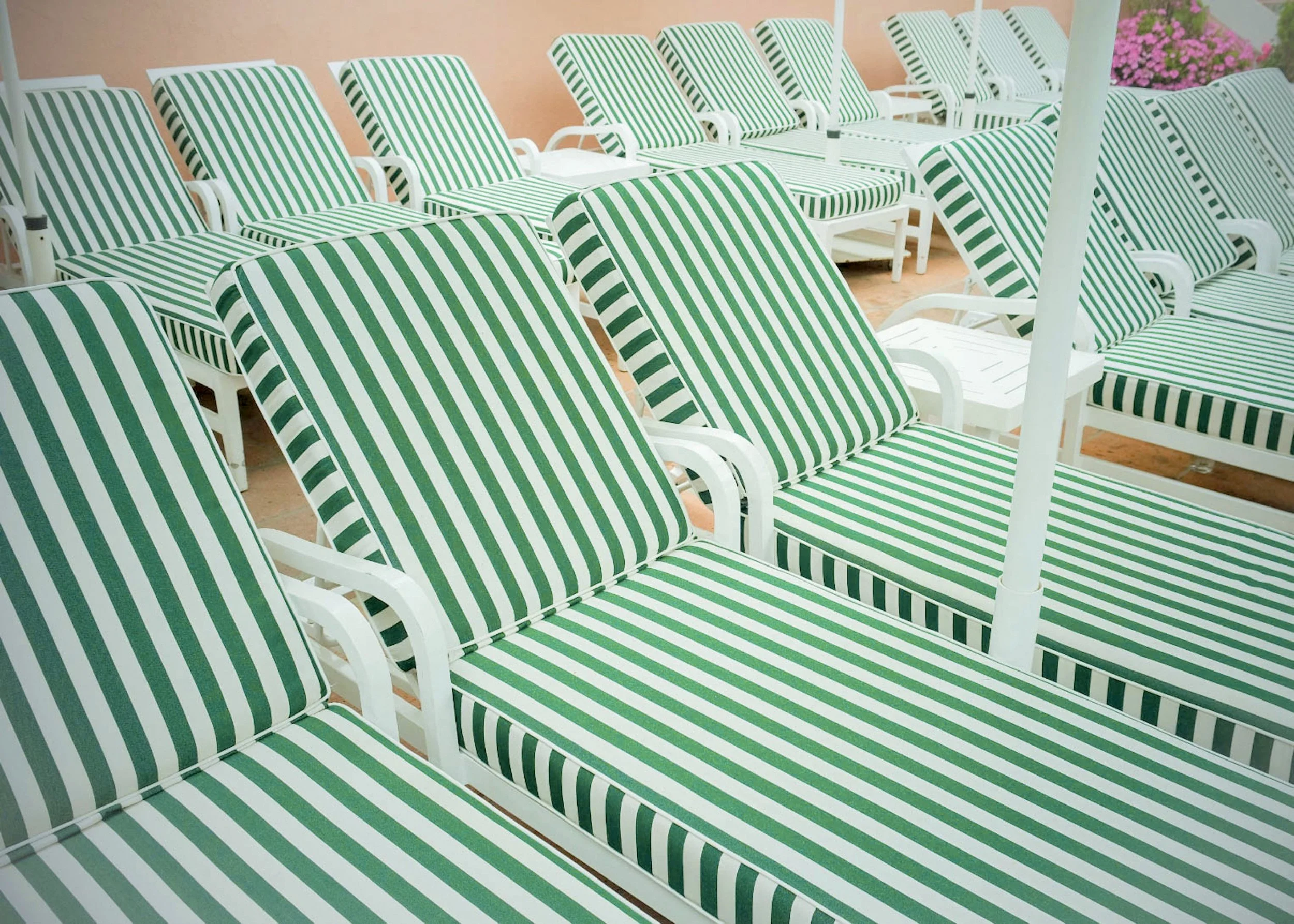 Multiple white lounge chairs with green and white striped cushions arranged outdoors near a pink wall and pink flowers in the background.