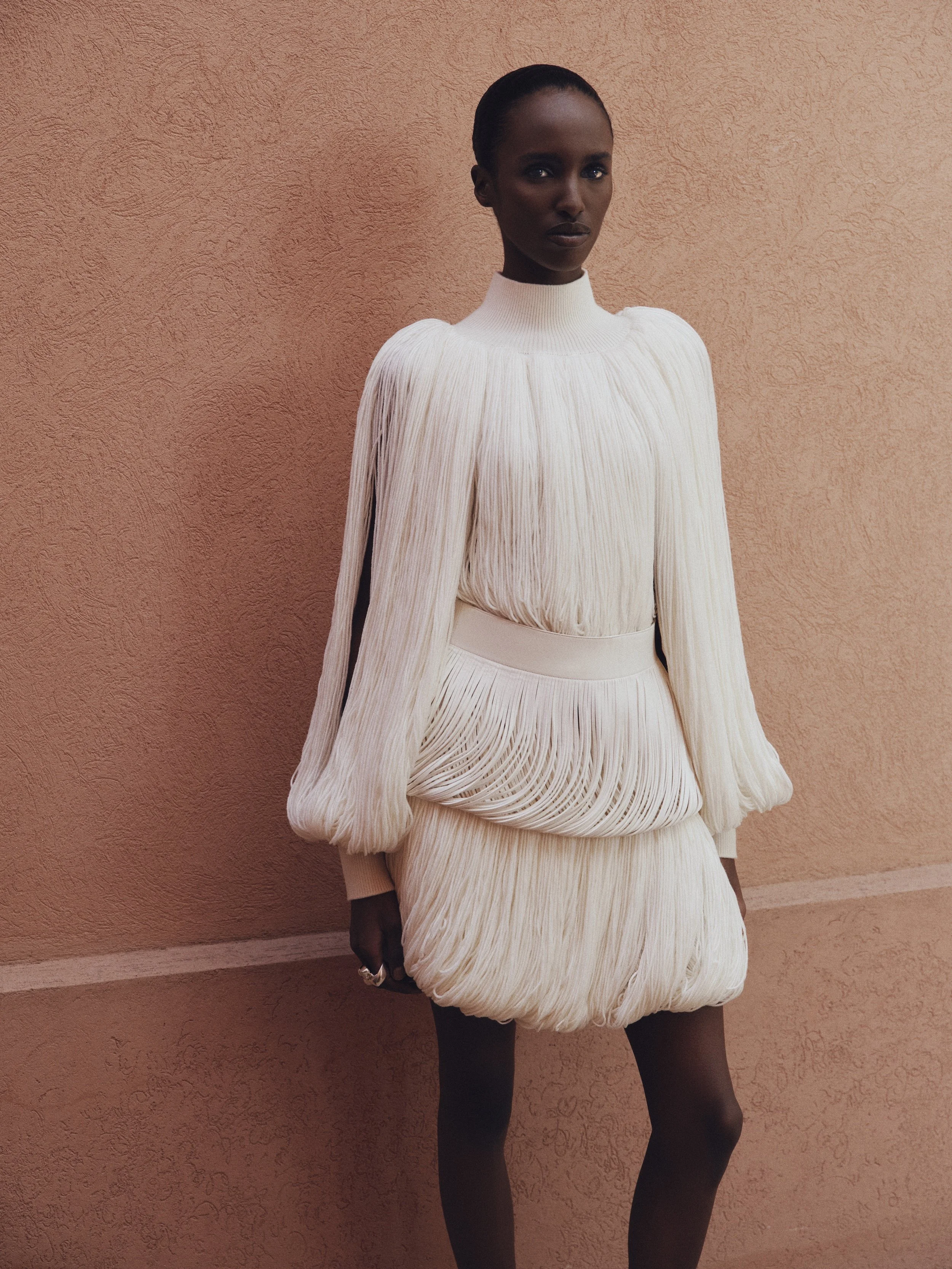 A woman in a white pleated dress with long sleeves and a high neckline standing against a peach-colored textured wall.