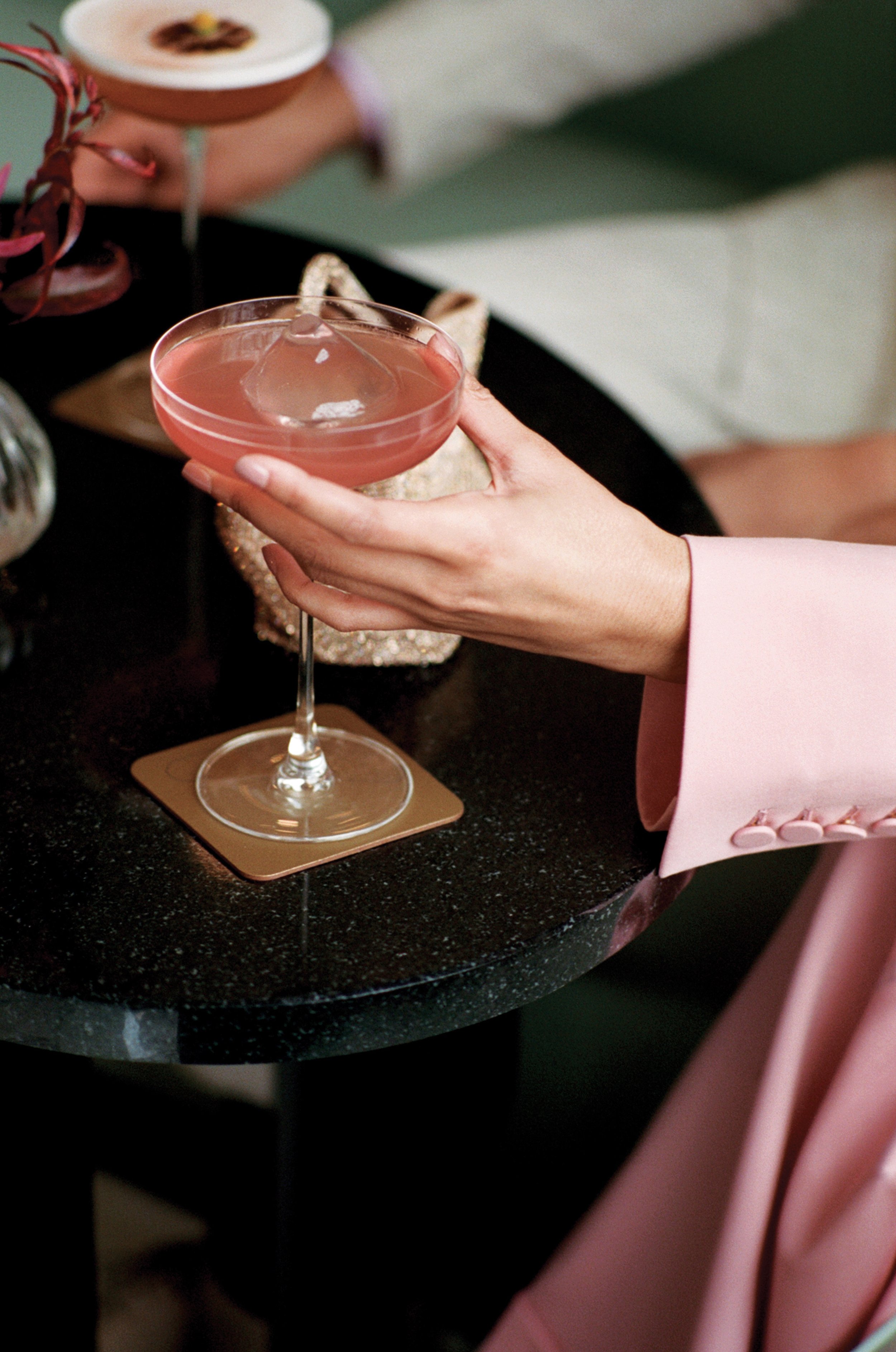 A person holding a pink cocktail garnished with a citrus twist, with a second cocktail visible in the background on a black marble table.