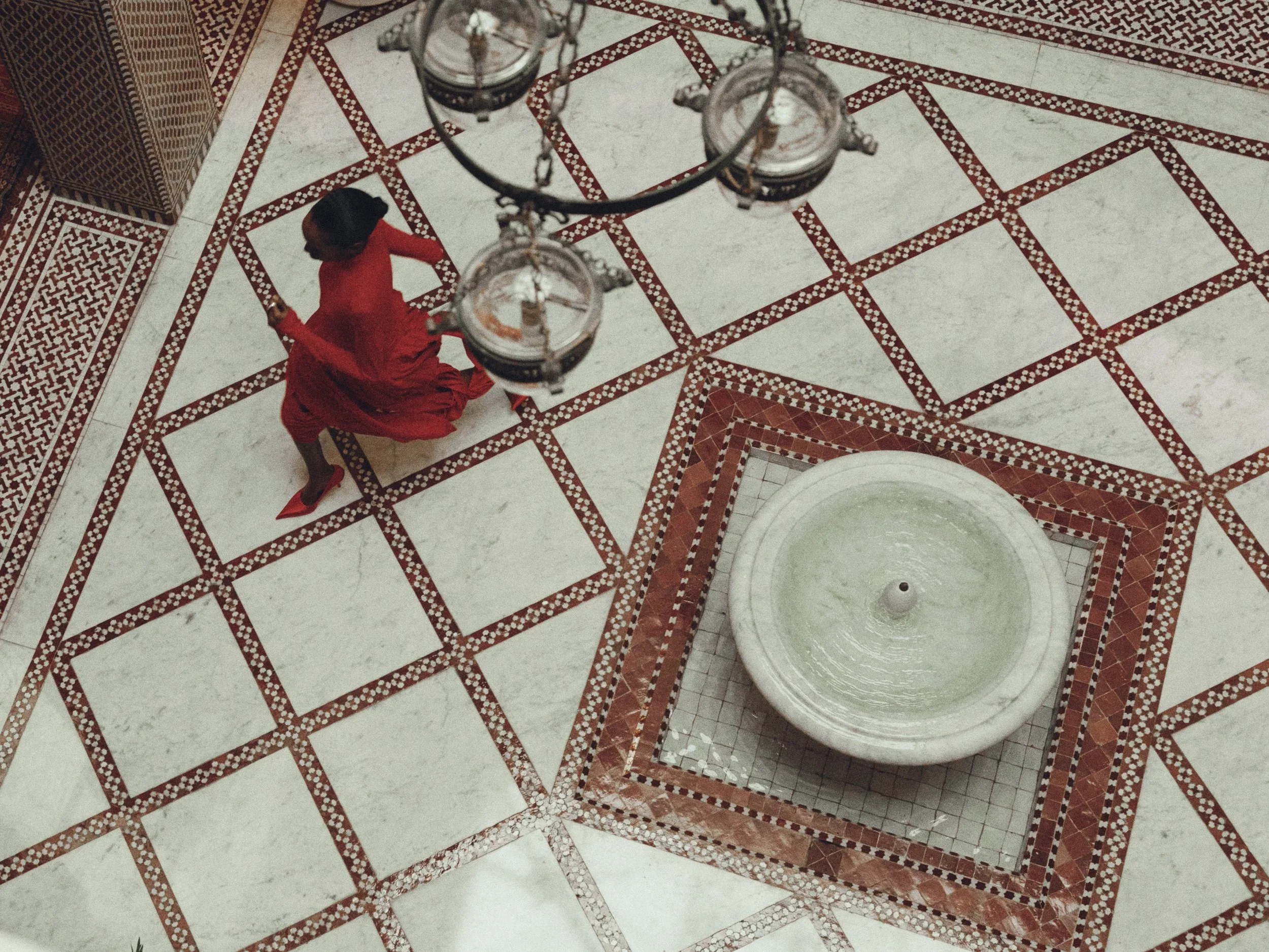 A woman dressed in a red outfit walking near marble and red patterned tile flooring, with a large white marble fountain and a decorative hanging chandelier overhead.