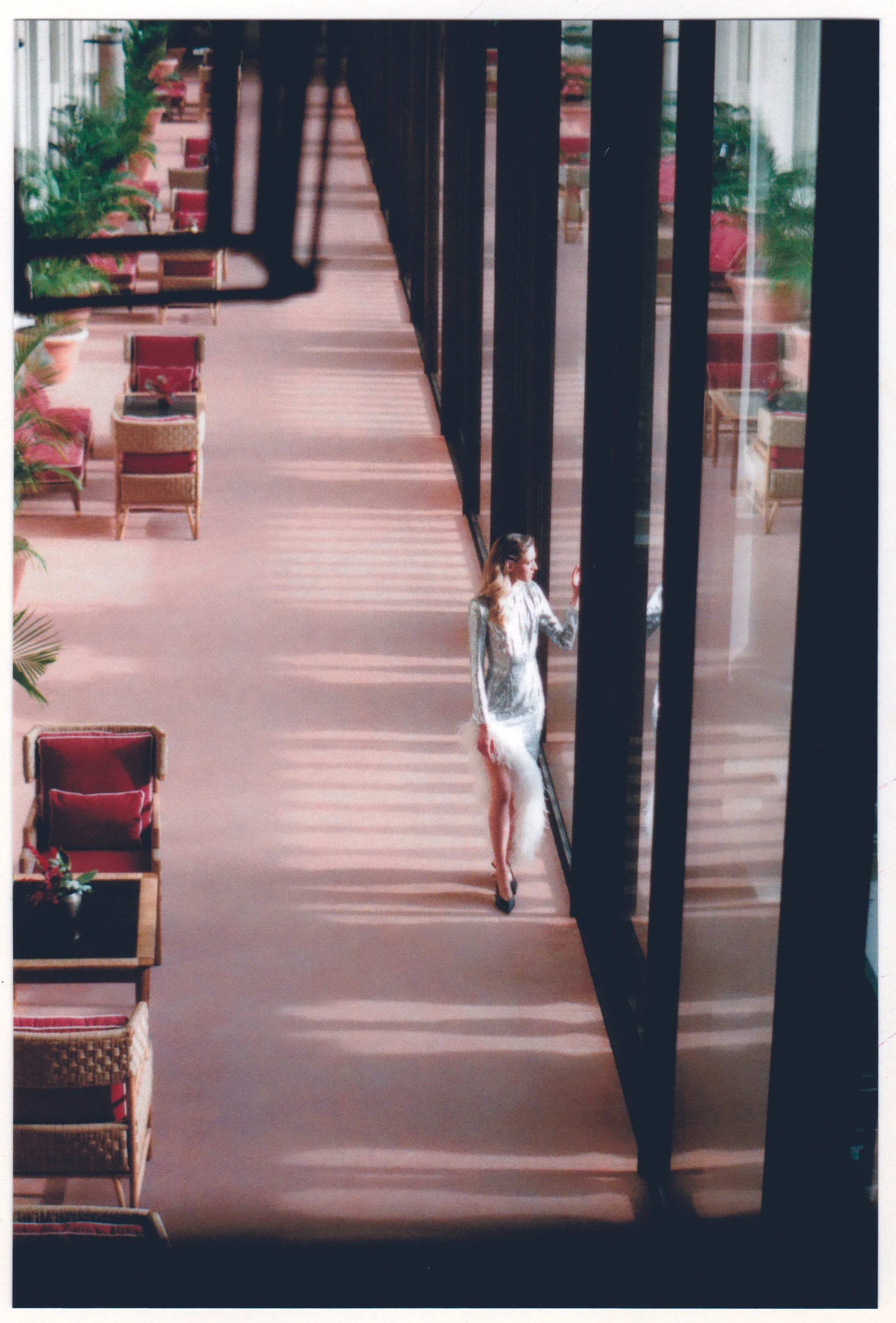A woman with long blonde hair standing by glass doors, wearing a sheer dress with floral patterns, dark high heels, inside a room with outdoor chairs and plants visible through the doors.