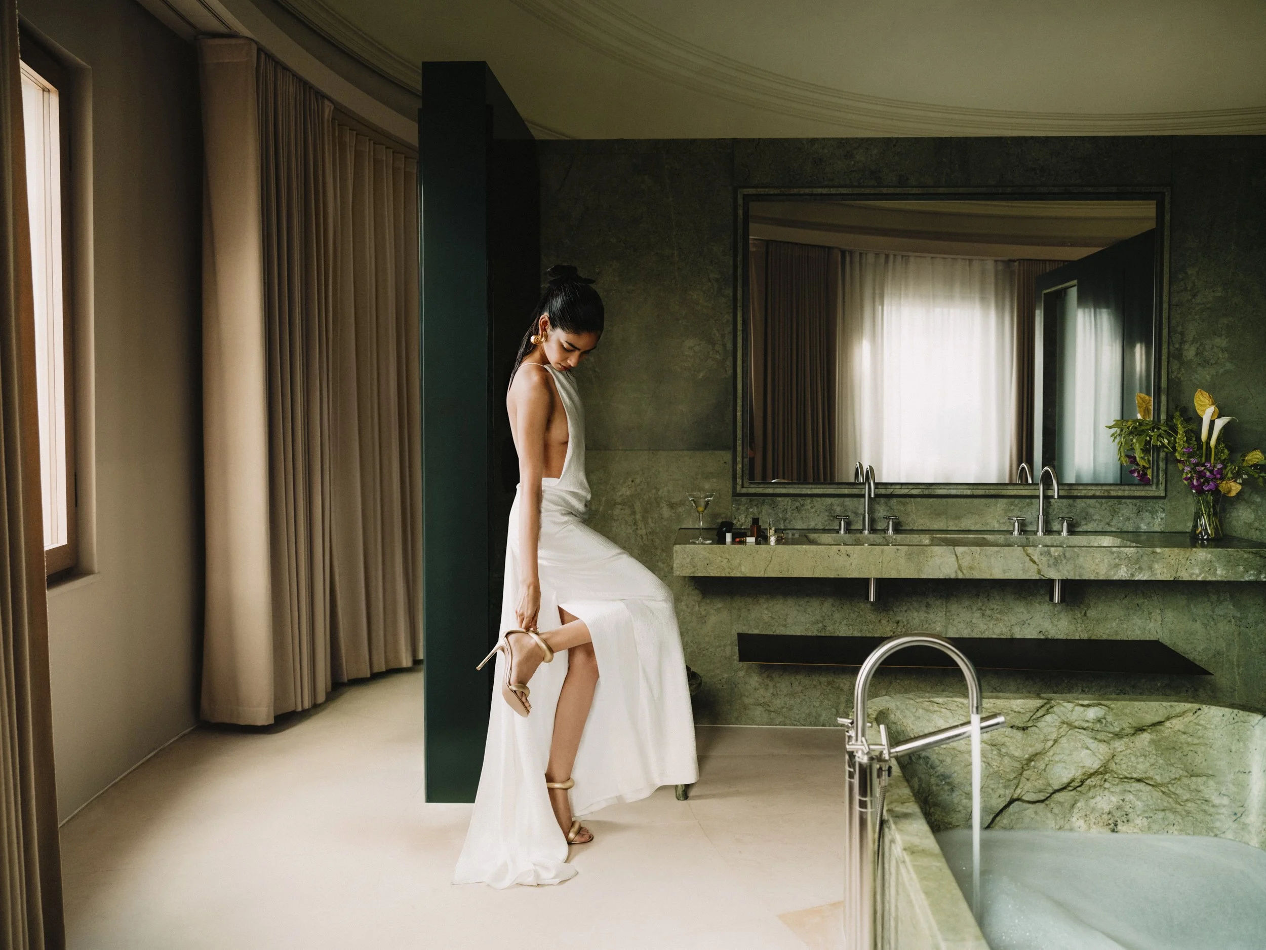 A woman in a white dress is standing in a modern bathroom, adjusting her high-heeled shoe. The bathroom features a large mirror, green marble countertops, and a window with curtains, sunlight filtering through.