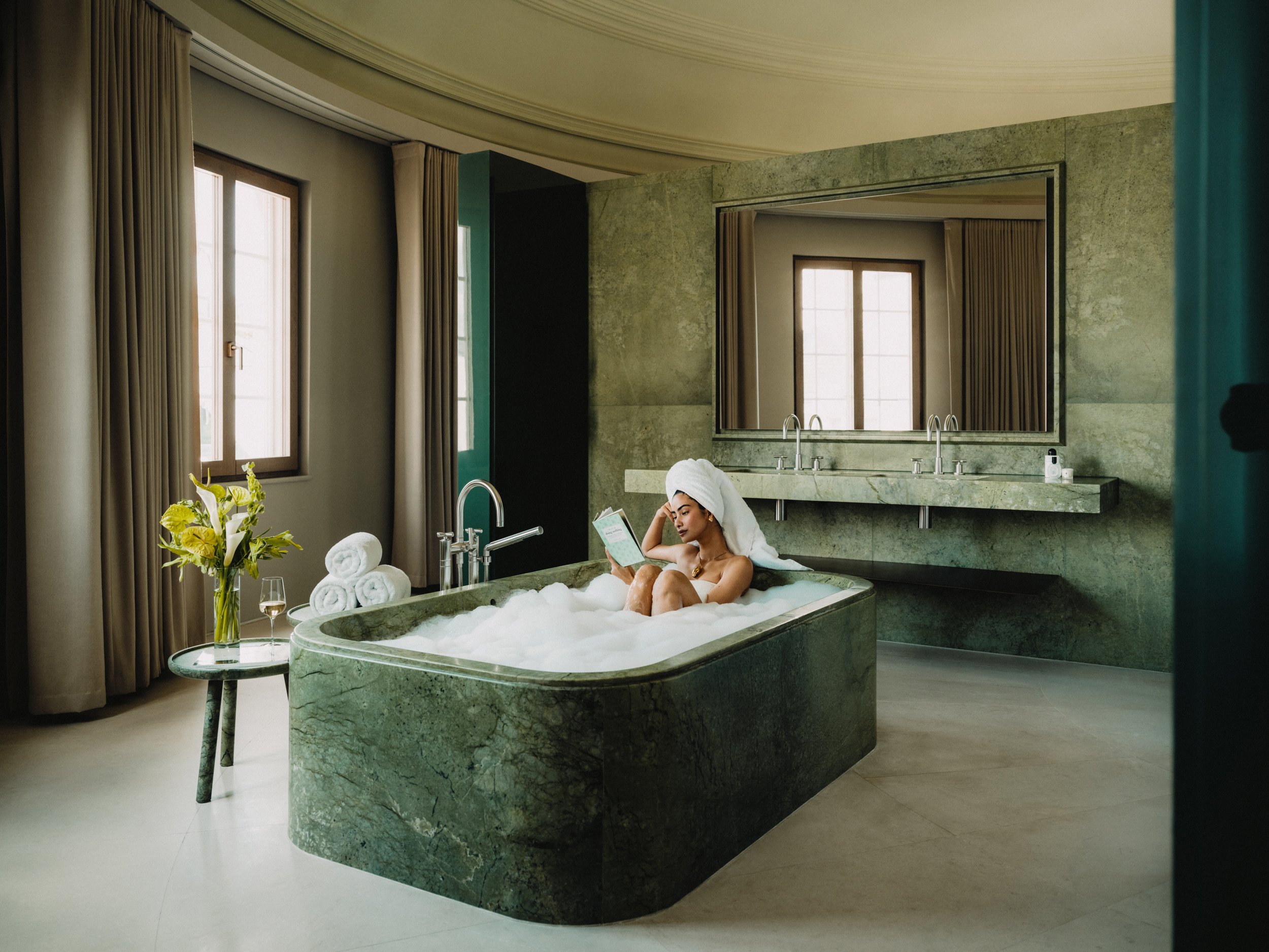 A woman with a towel on her head relaxing in a bathtub filled with bubbles in a luxurious bathroom, with a large mirror and windows letting in natural light.