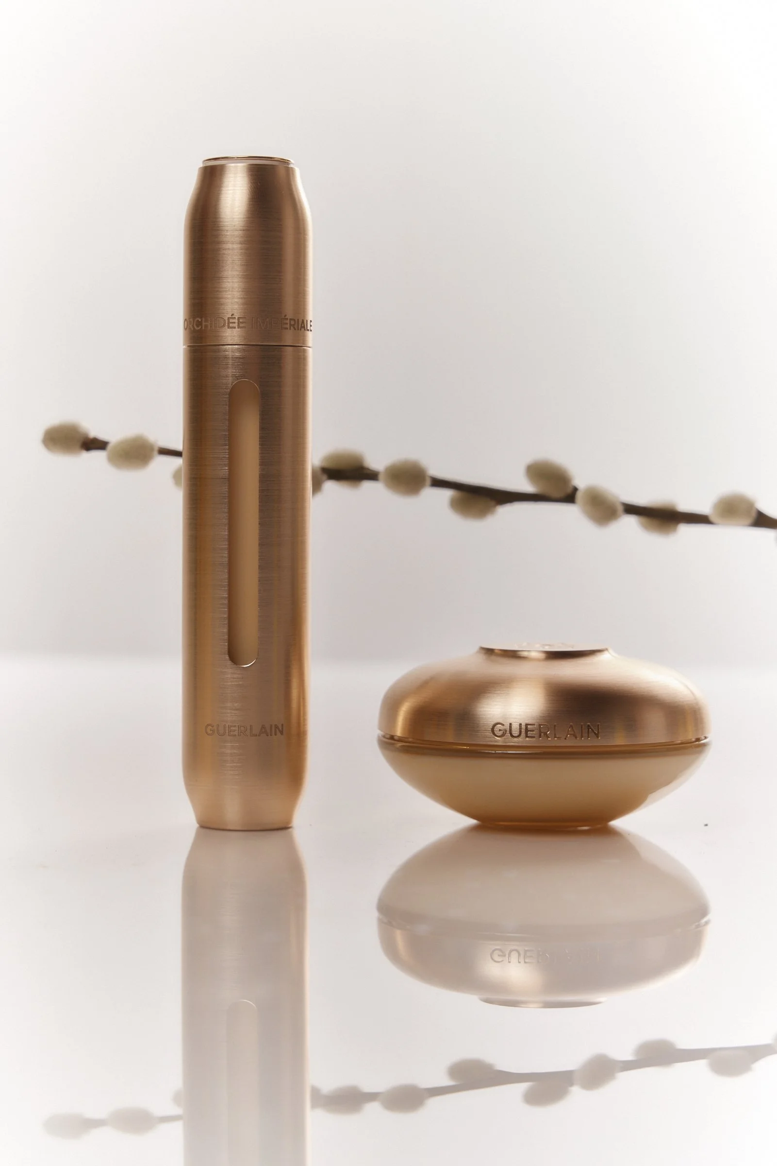Guerlain skincare products with a branch of pussy willow in the background, all on a reflective white surface.