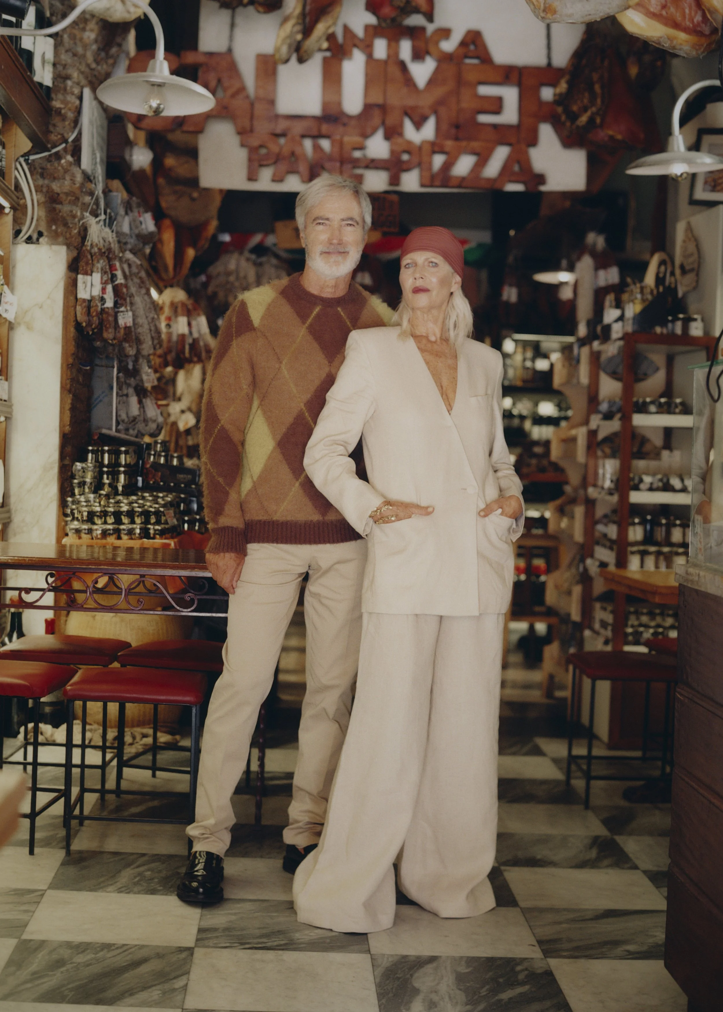 Older man and woman standing inside a store with food items and a sign that says 'Antica Pizzeria' in the background.