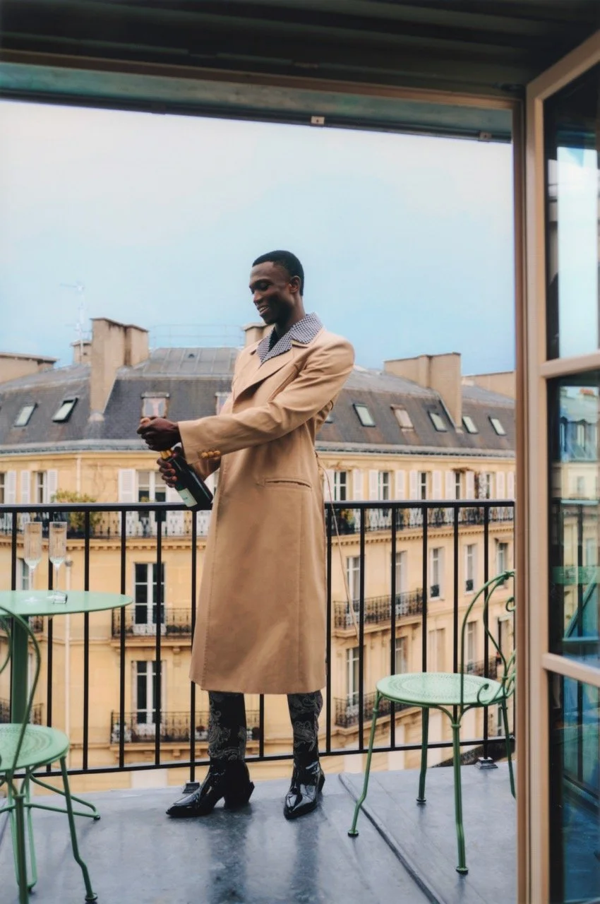 A person standing on a balcony holding a bottle, wearing a beige trench coat, patterned pants, and black boots, with a cityscape of European-style buildings in the background.