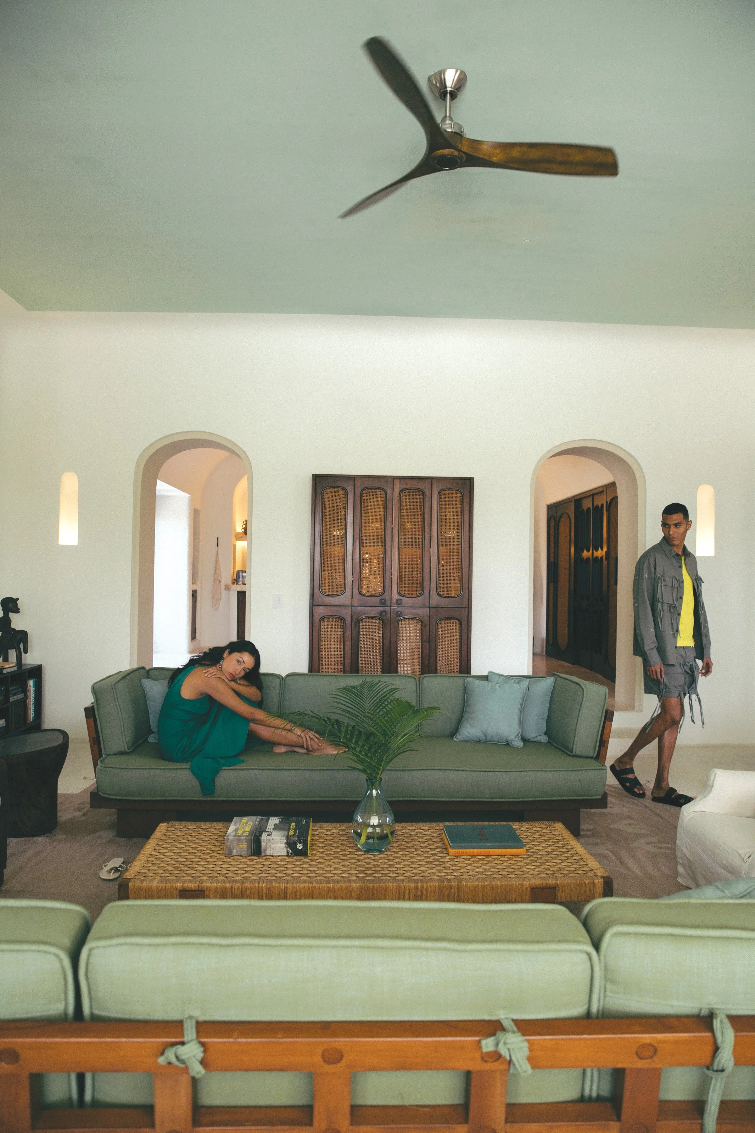 A woman with long dark hair wearing a teal dress stretches on a green sofa in a spacious living room, while a young man in a gray jacket, yellow shirt, shorts, and sandals walks by near a doorway. The room has a ceiling fan, wooden cabinets, arched d