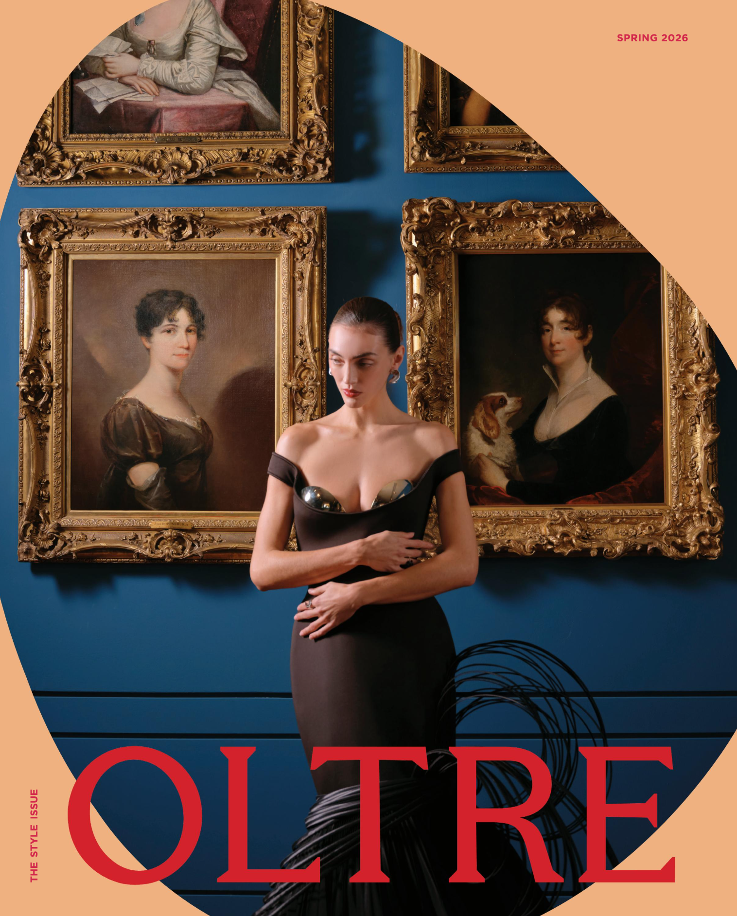 A woman with dark hair styled in a sleek bun, wearing a form-fitting black dress with metallic embellishments on the bodice, standing in front of a blue wall with four large ornate gold-framed paintings, and part of a red text that reads 'OU' and 'RE