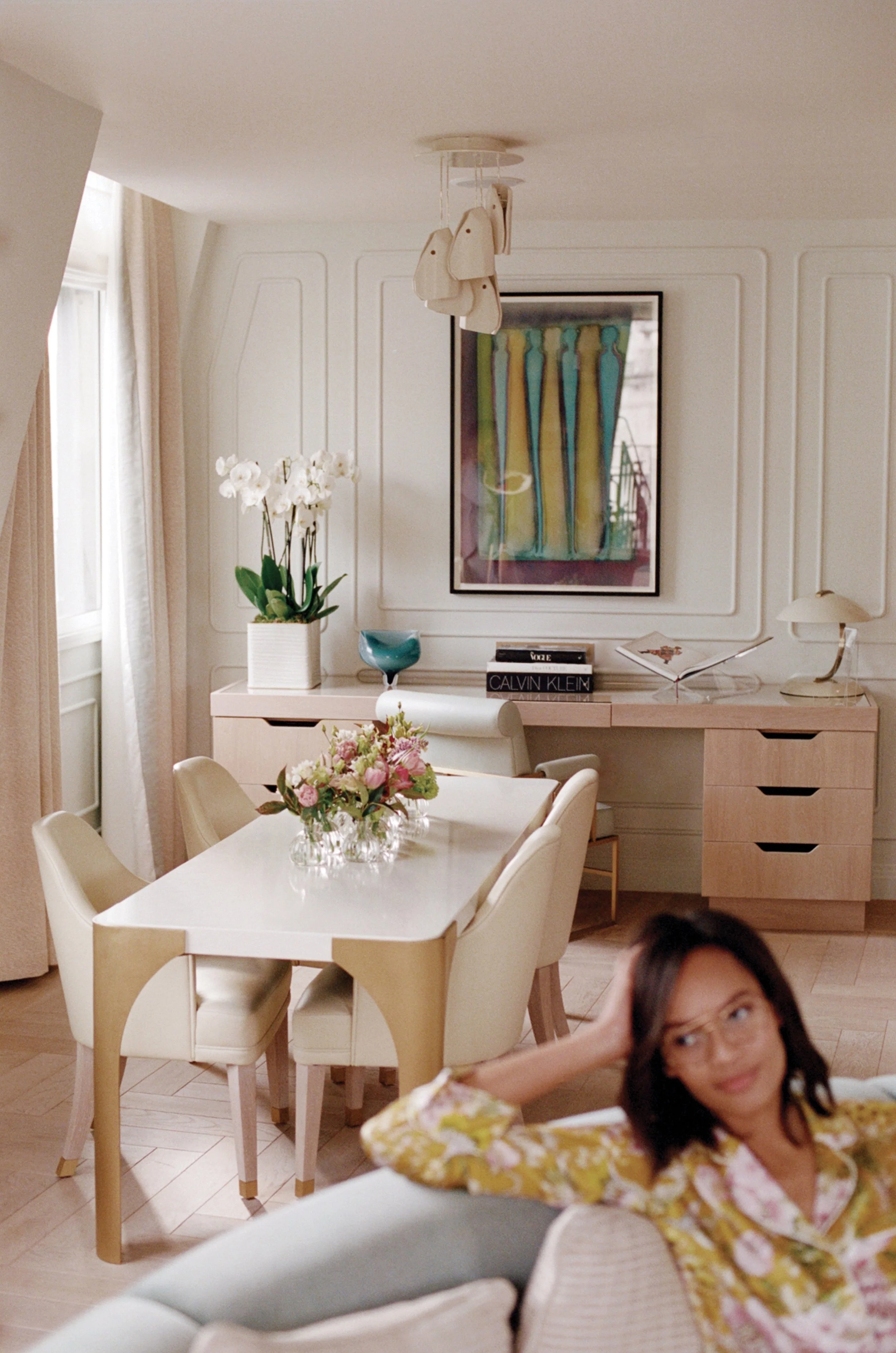 A woman in a floral yellow and pink dress lounging on a chair in a bright, modern dining room with white walls, a white dining table with a floral centerpiece, white chairs, a sideboard with decorations including a large orchid, a colorful abstract p