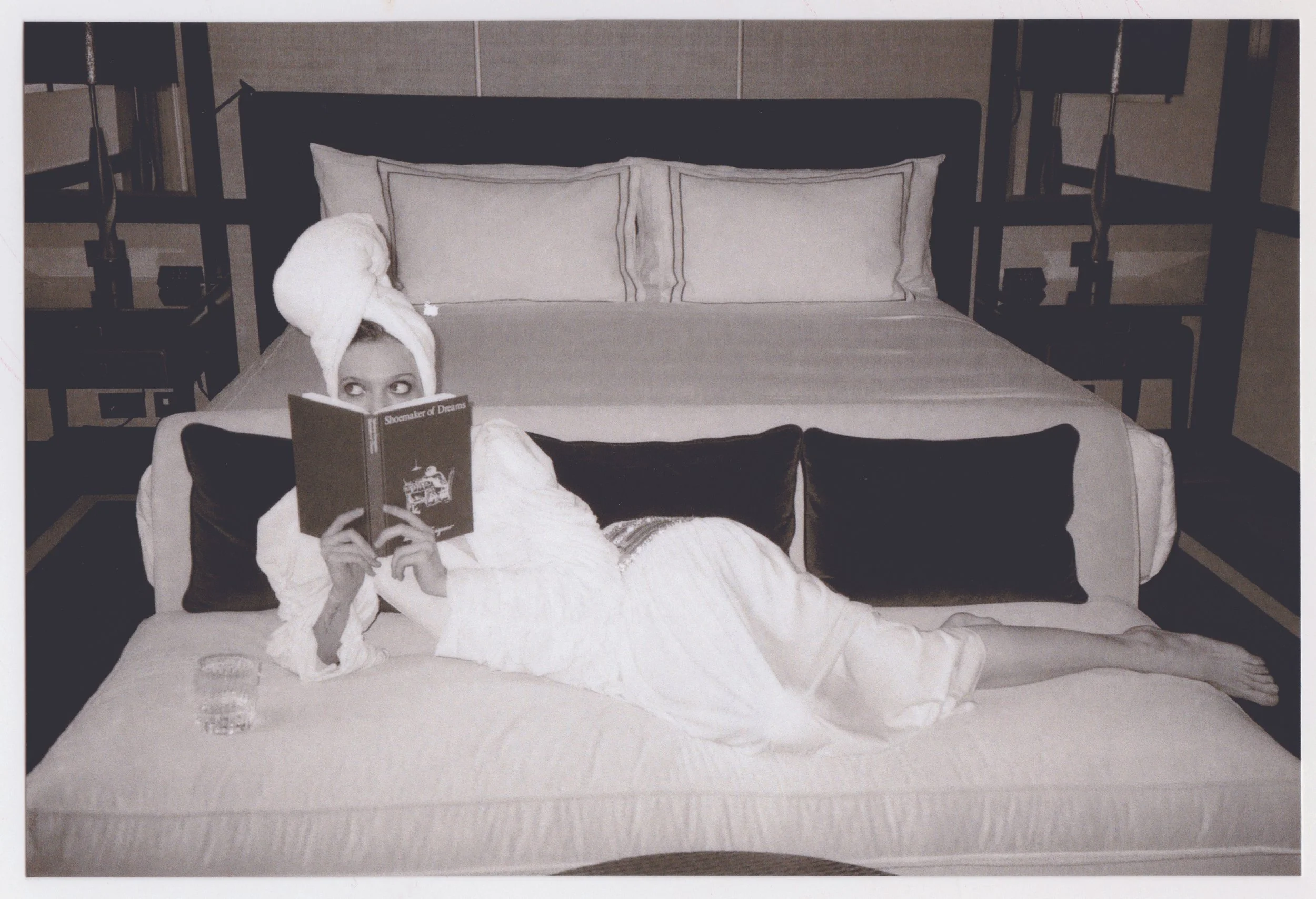 A person lying on a bed with a towel wrapped around their head, reading a book titled 'Shoemaker of Dreams,' in a hotel room with a large headboard, three pillows, and side tables with lamps.