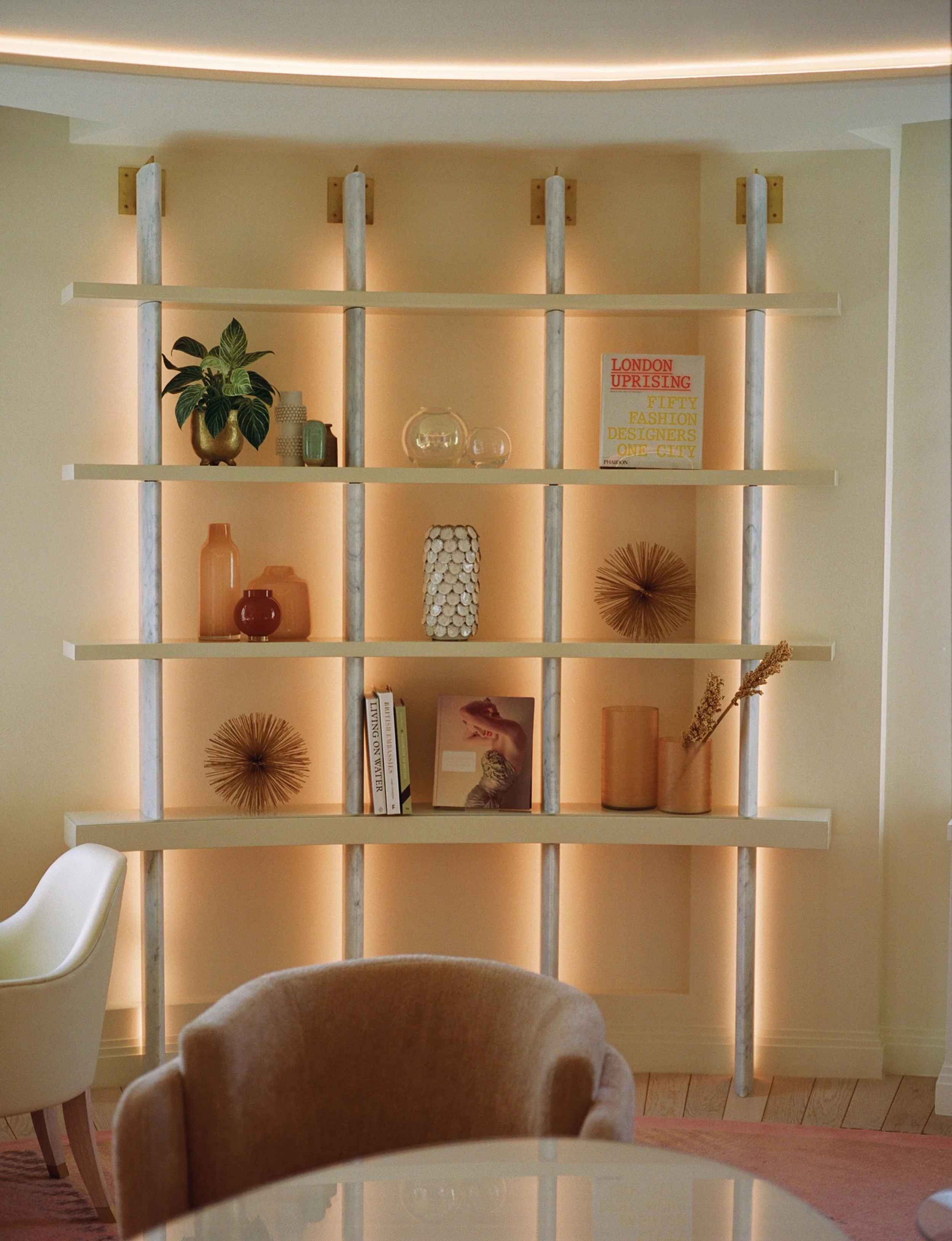 A modern interior space with a shelving unit against a beige wall illuminated by warm lighting. The shelves display decorative items including books, vases, glass bowls, and art pieces. Part of a white chair and a brown upholstered chair are visible 