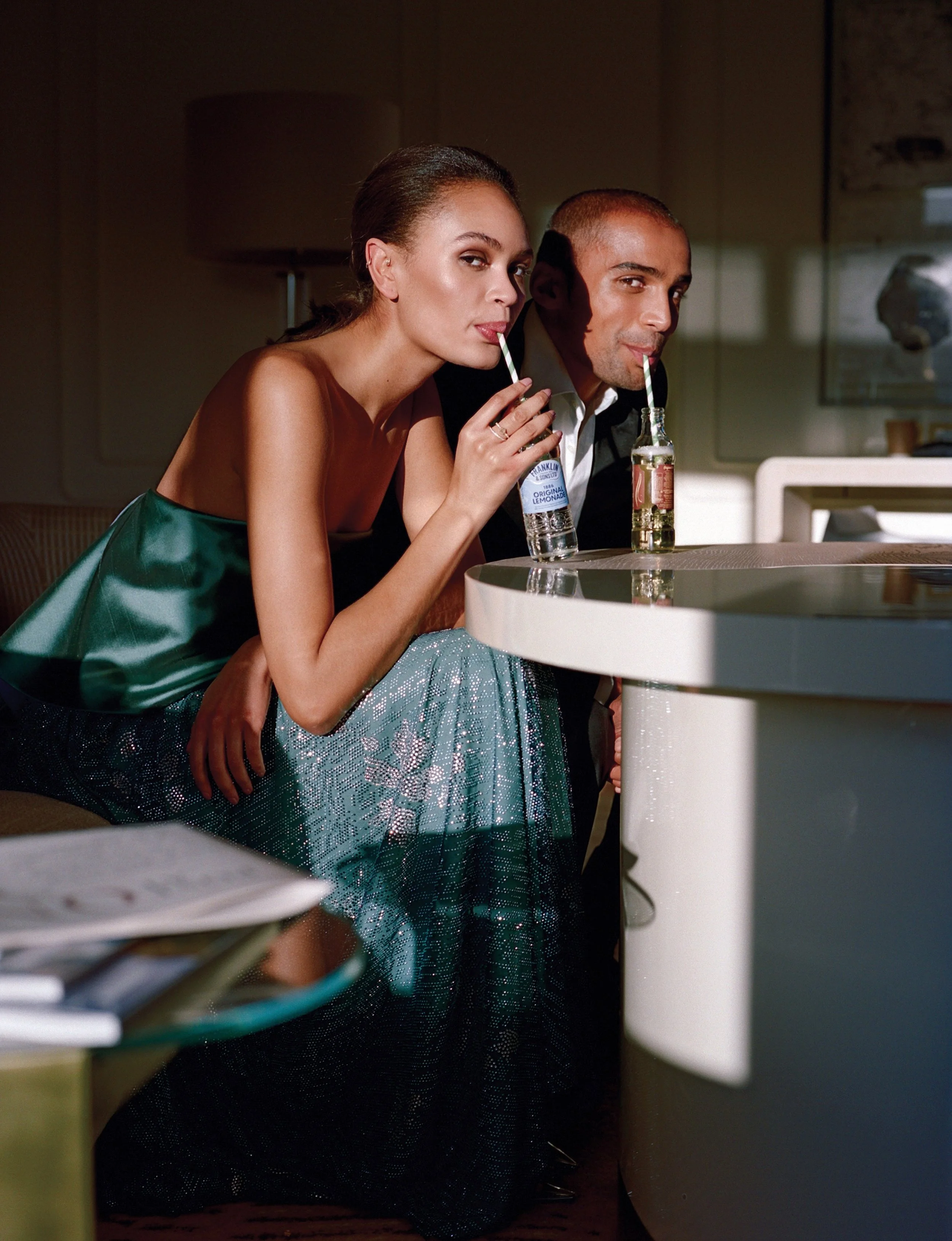 A woman and a man sitting at a table, each drinking from glass bottles with straws, in a modern indoor setting.
