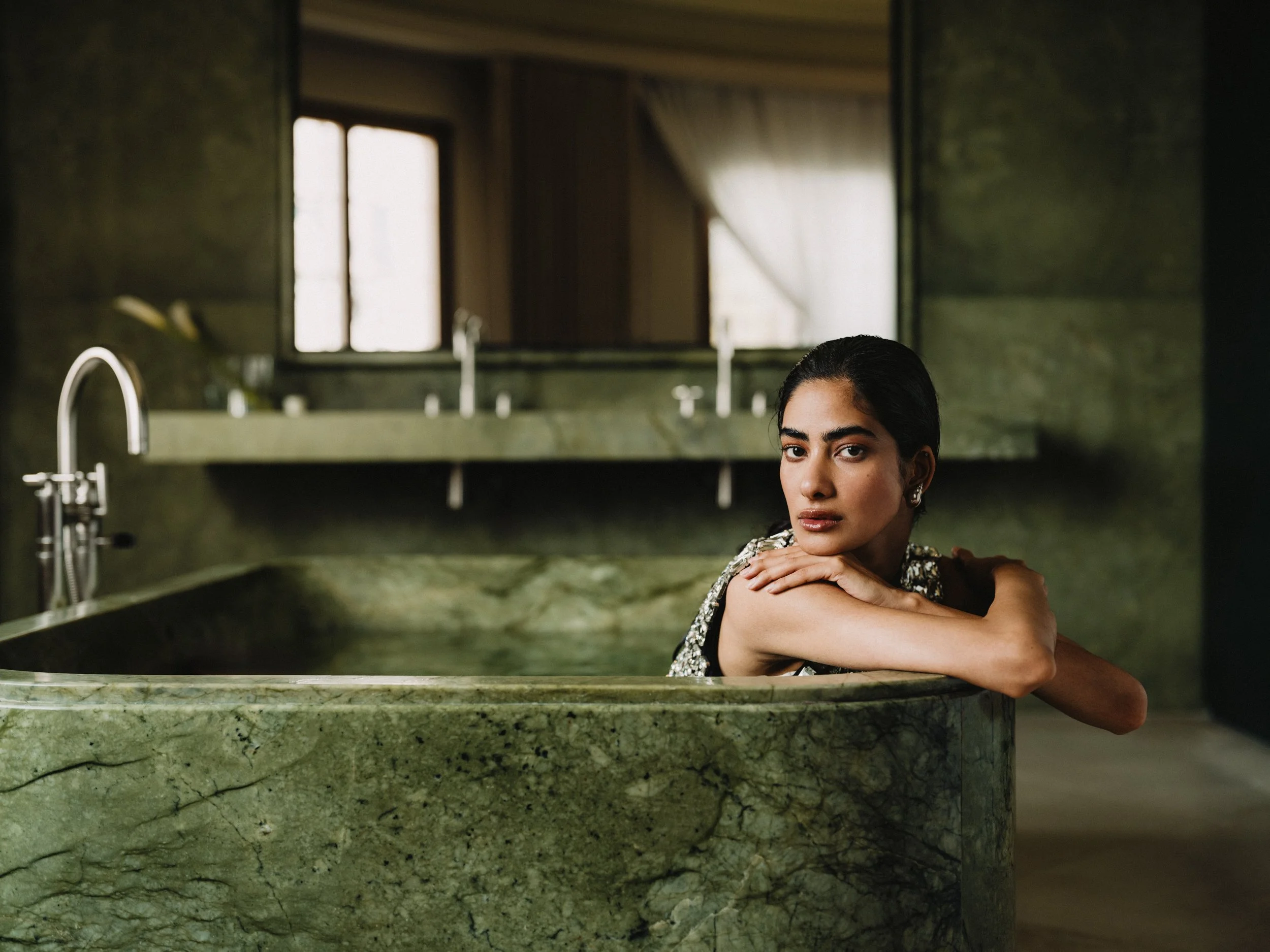 A woman with dark hair and a floral dress leaning on a green marble bathtub in a bathroom, with a window in the background.