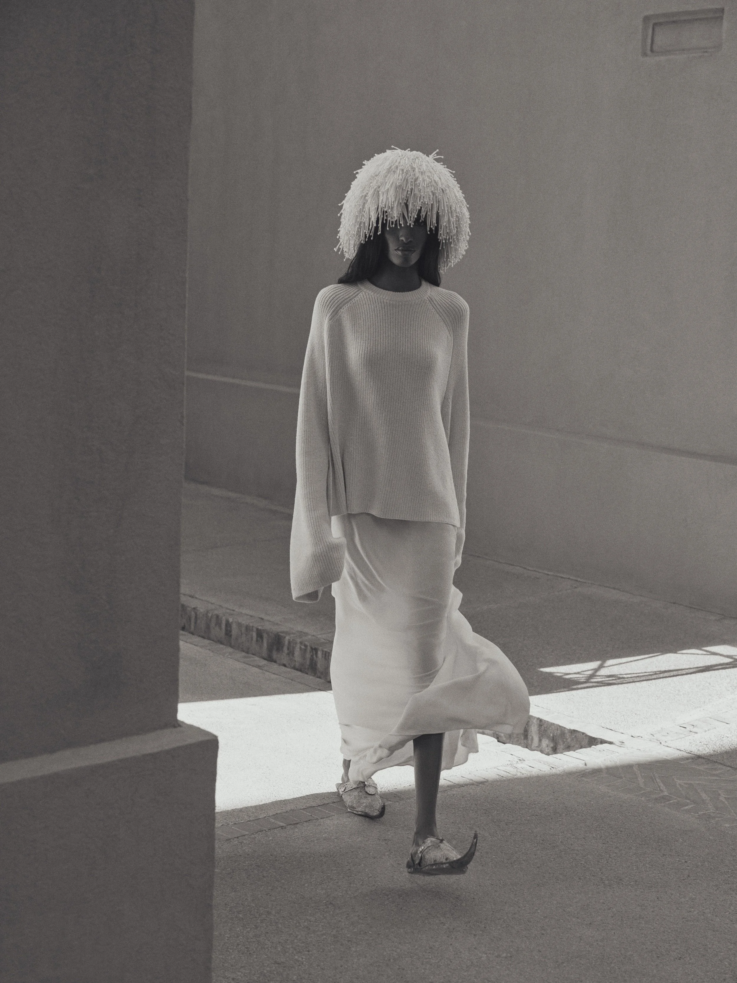 A woman with dark hair wearing a textured, long-sleeved, off-white sweater and a white skirt walking outdoors in sunlight, with a large, elaborate hat made of textured material covering her eyes, against a plain, minimalist background.