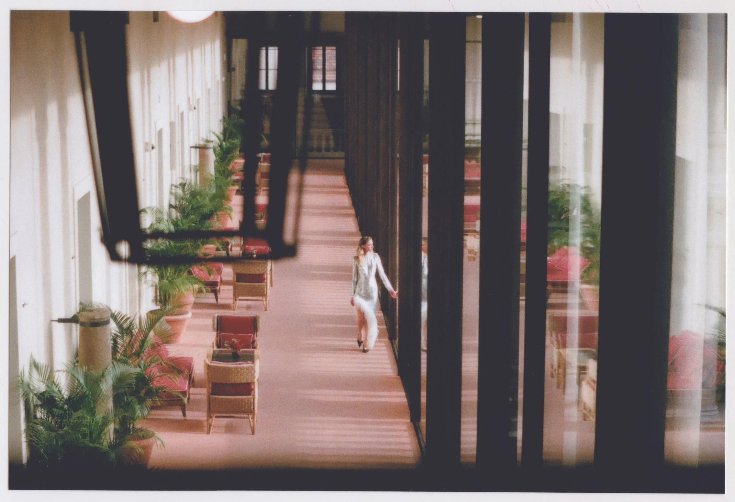A woman in white pants and a blazer walking in a corridor decorated with potted plants and chairs, framed by large black vertical pillars.