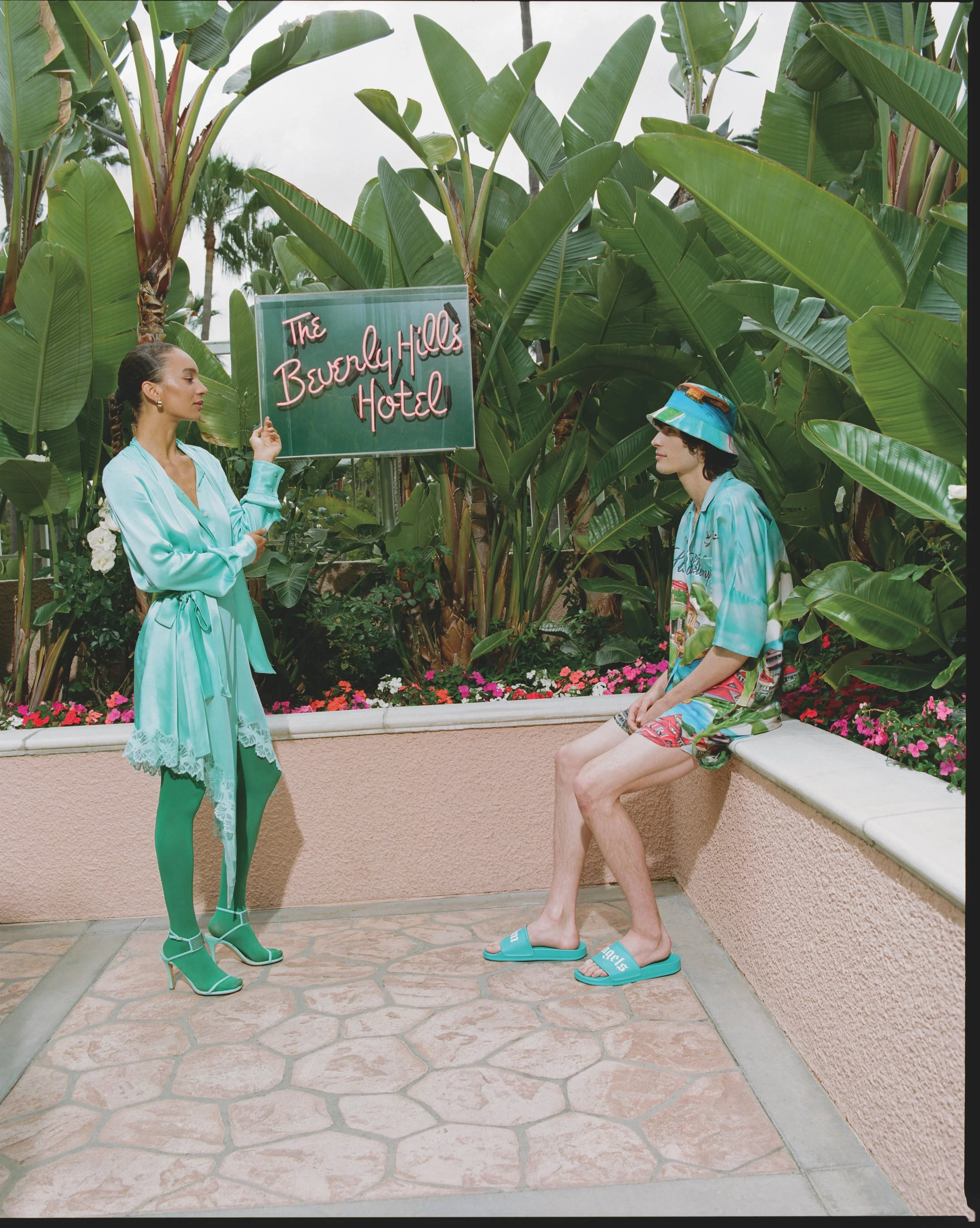 A woman with brown hair pulled back in a braid, dressed in a teal satin robe, is standing on the left, near a green sign that reads 'The Beverly Hills Hotel'. She is looking at a man who is sitting on a ledge, wearing a colorful hat, patterned shirt,