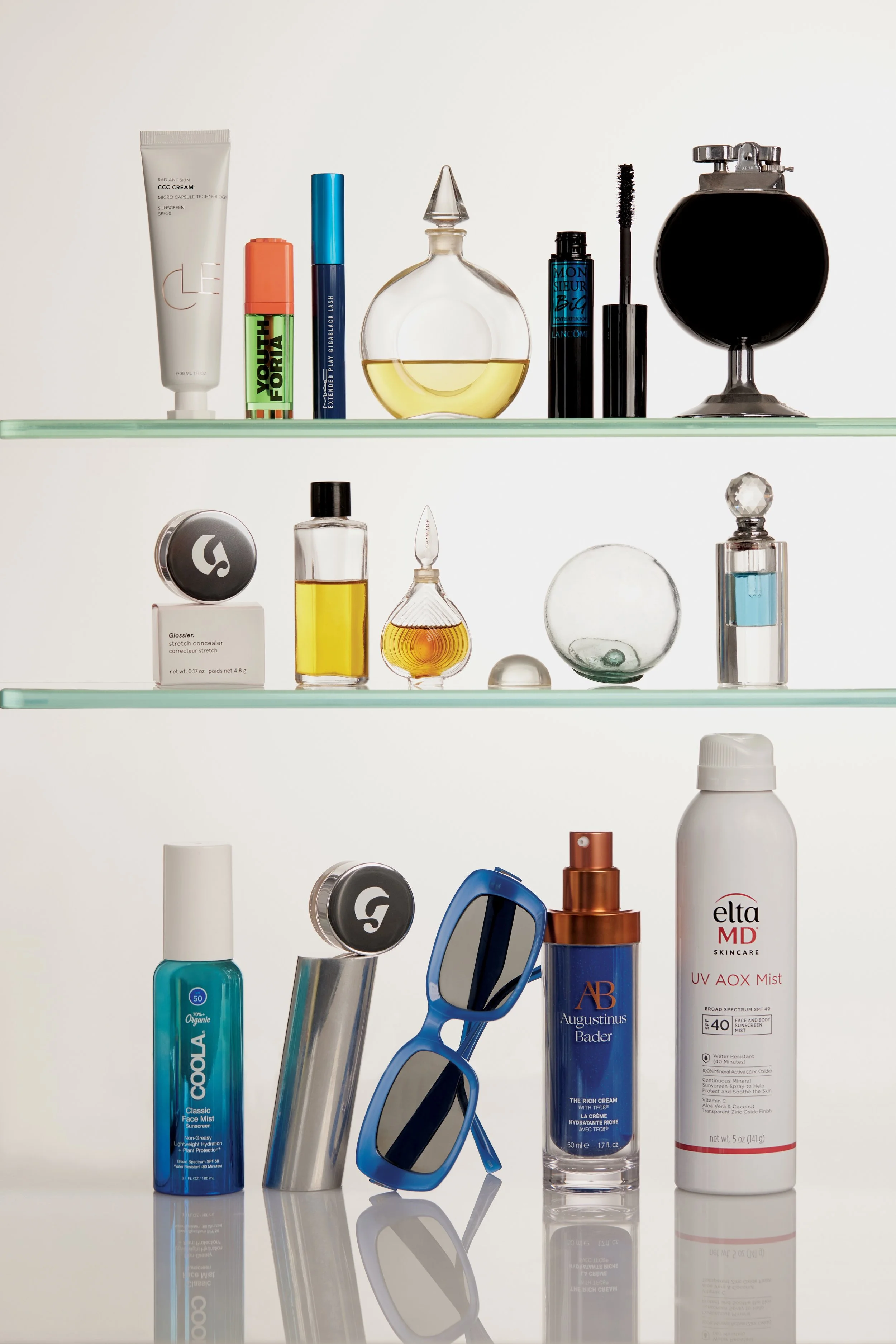 A collection of skincare and beauty products displayed on glass shelves, including lotions, sprays, a perfume bottle, makeup, sunglasses, and sunscreen.