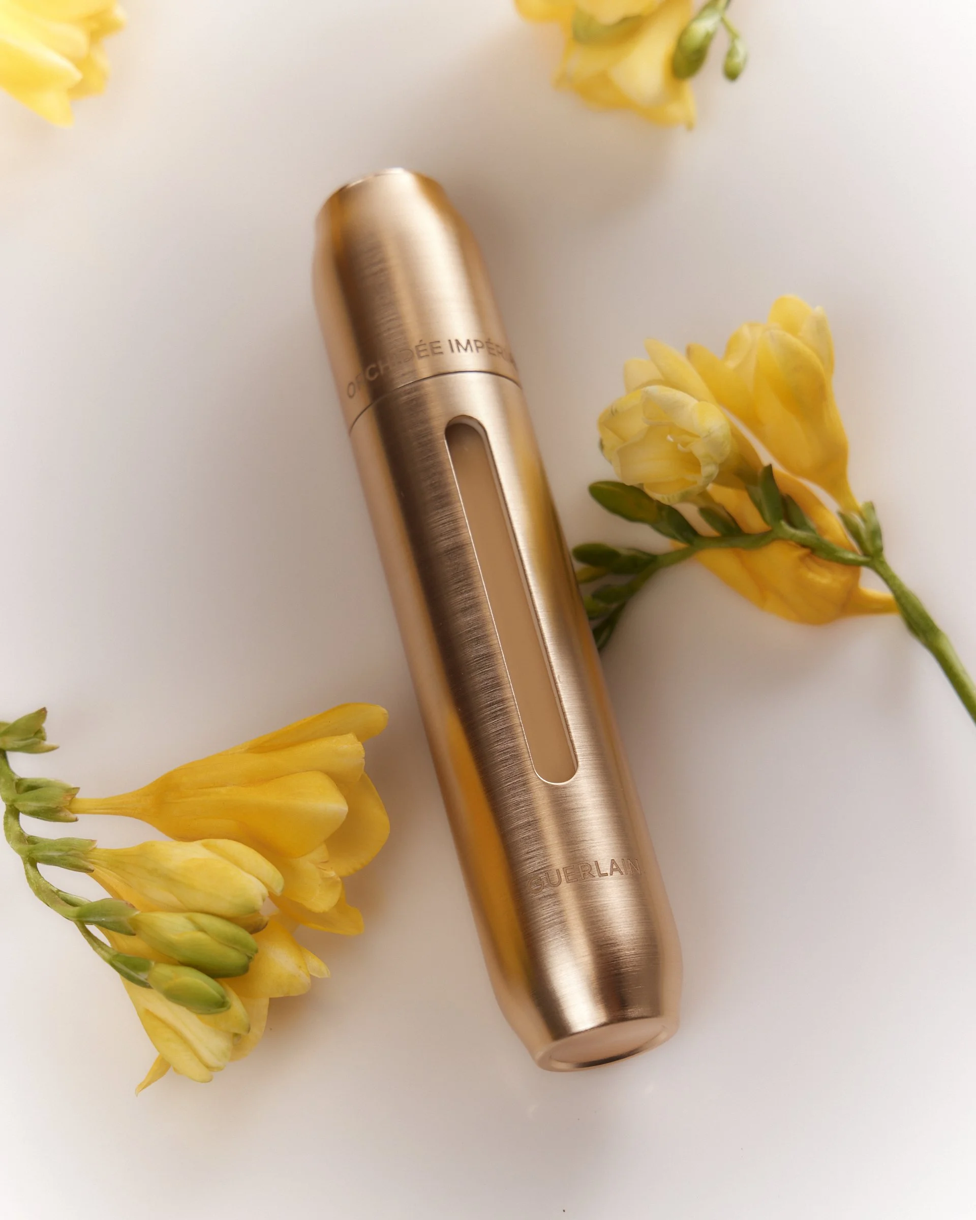 Gold-colored skincare serum bottle labeled 'Guerlain' surrounded by yellow freesia flowers on a white background.