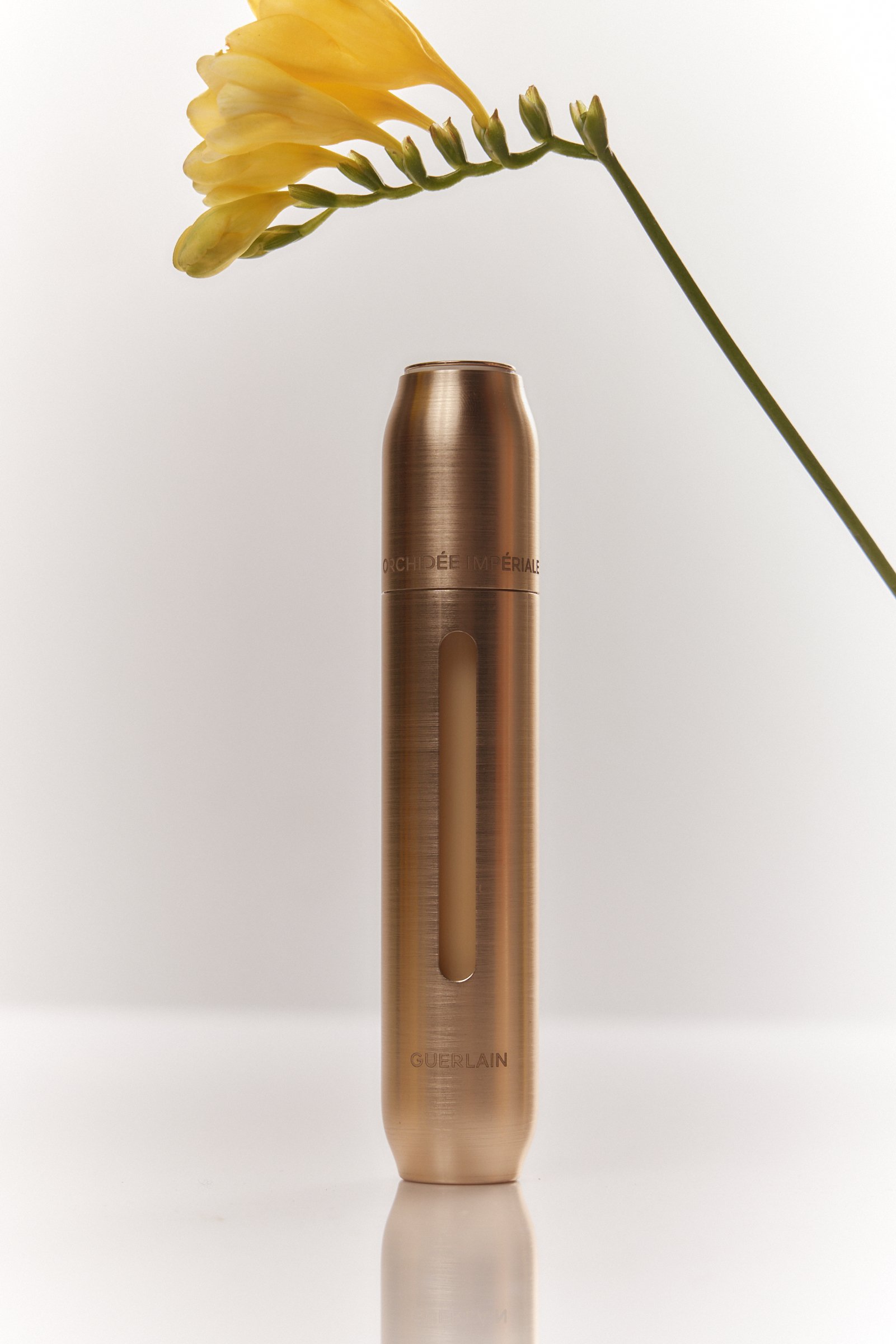 A rose gold cosmetic spray bottle with the words "Guerlain" on it, standing upright on a reflective surface, with a white flower with yellow petals extending from its top.