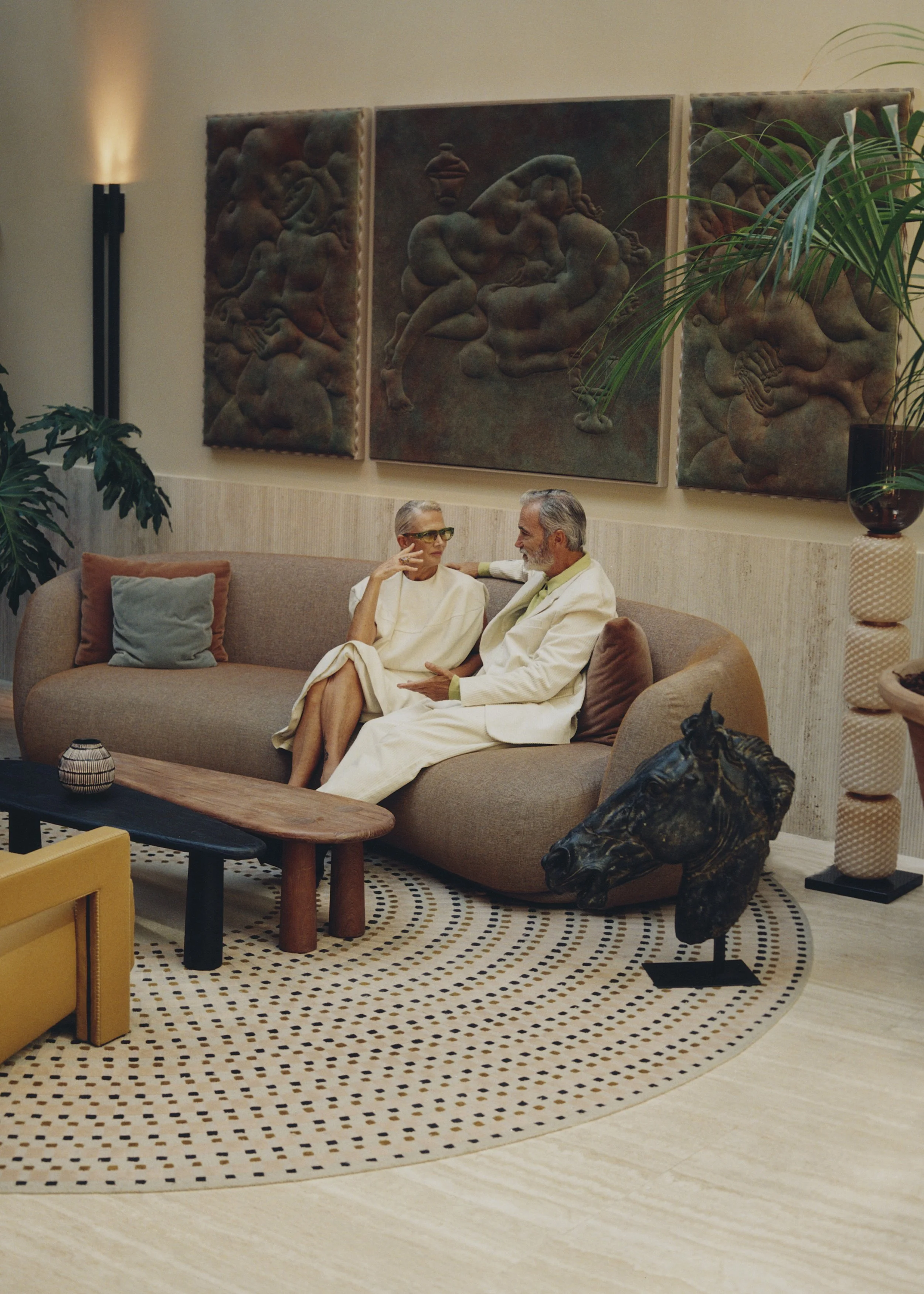 A mid-century modern living room with a beige sofa, colorful pillows, a round patterned rug, and three wooden art panels on the wall. Two older adults are sitting on the sofa, engaging in conversation. A large potted plant is on the right, and part of a black horse head sculpture is visible in the foreground.