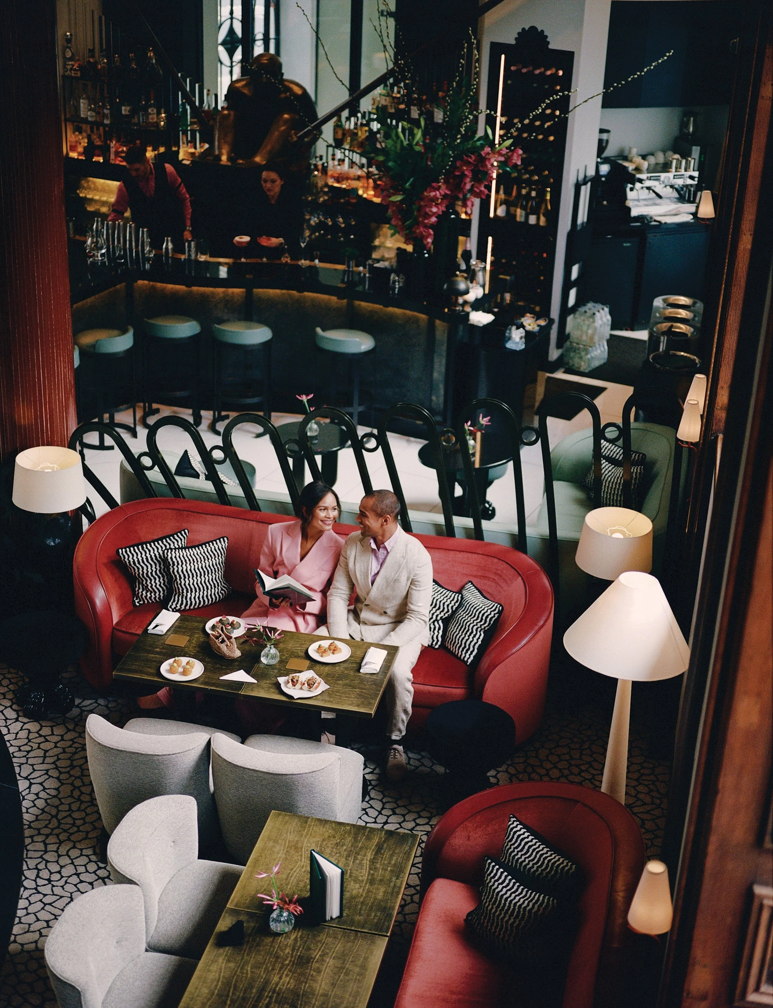 Couple sitting on a red sofa with black and white pillows, talking and smiling, in a cozy restaurant with white armchairs, small dining tables, and a bar area in the background.