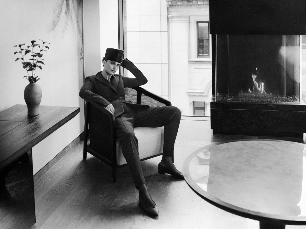 A black and white photo of a woman sitting on a chair in a modern, stylish room with a fireplace, a round table, and a vase with a plant, looking contemplative.