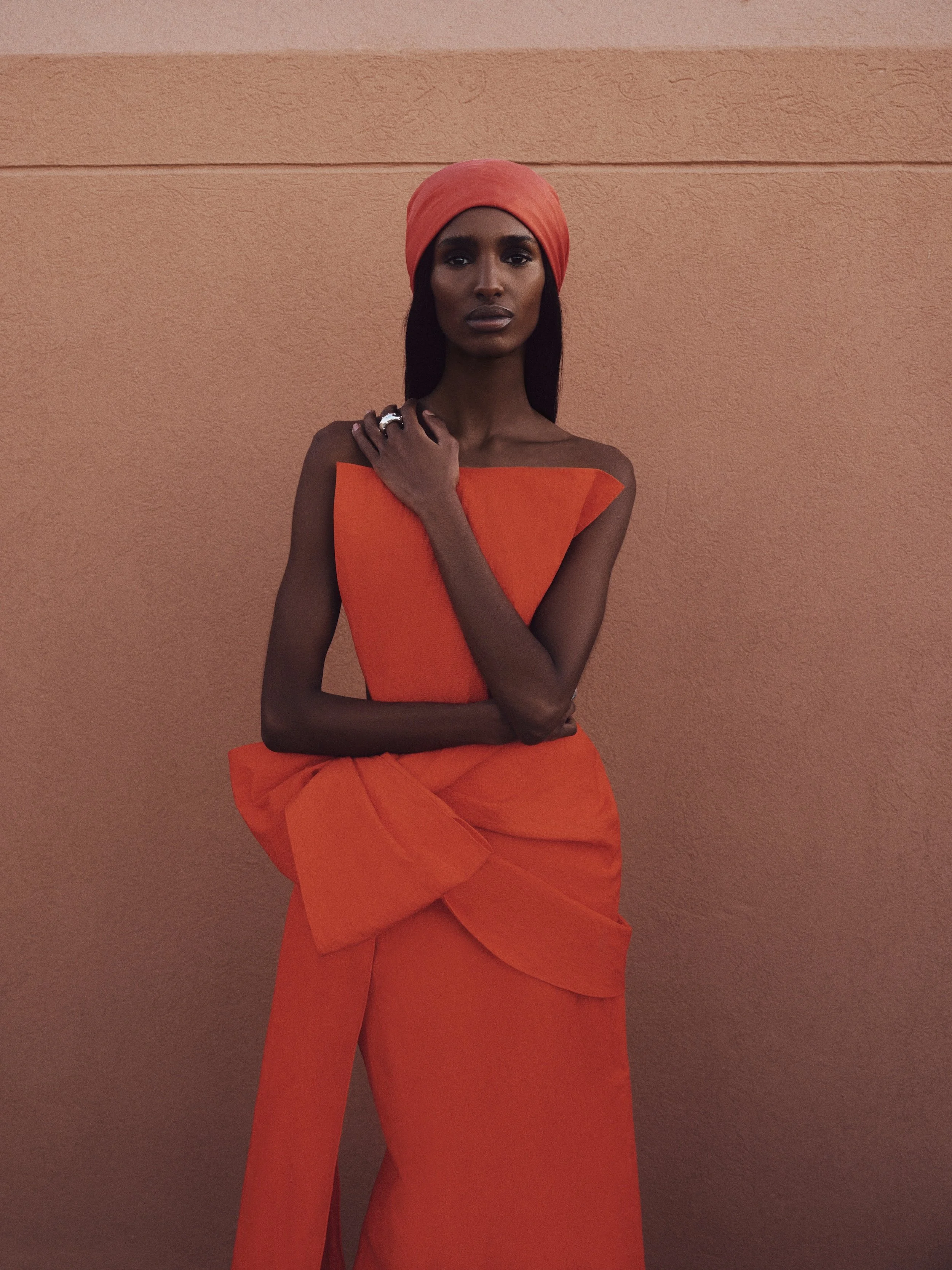 A woman wearing an orange head wrap and a matching orange off-the-shoulder dress, standing against a textured peach-colored wall.