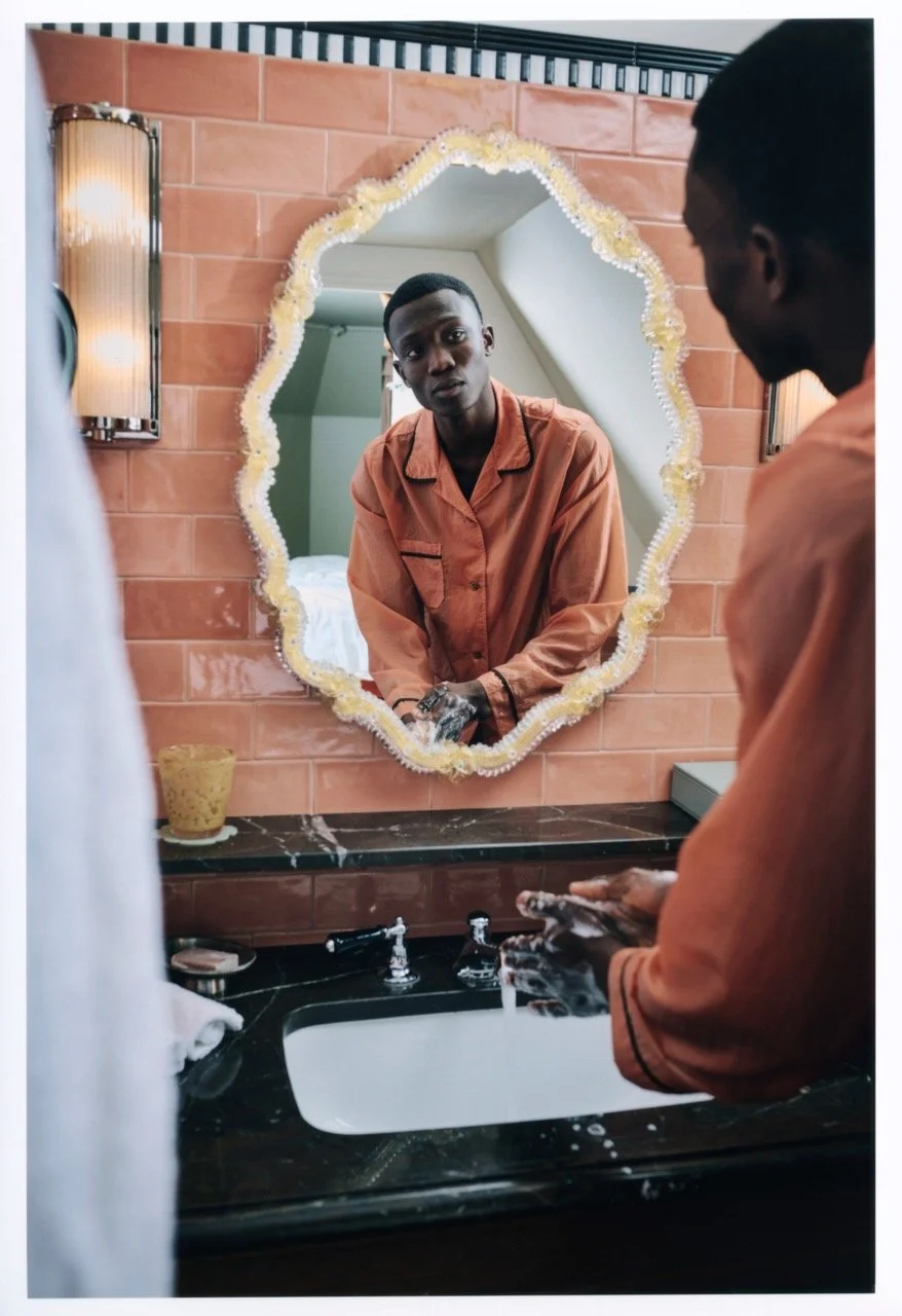A young man in an orange shirt looking at himself in a decorative mirror while washing his hands at a bathroom sink.