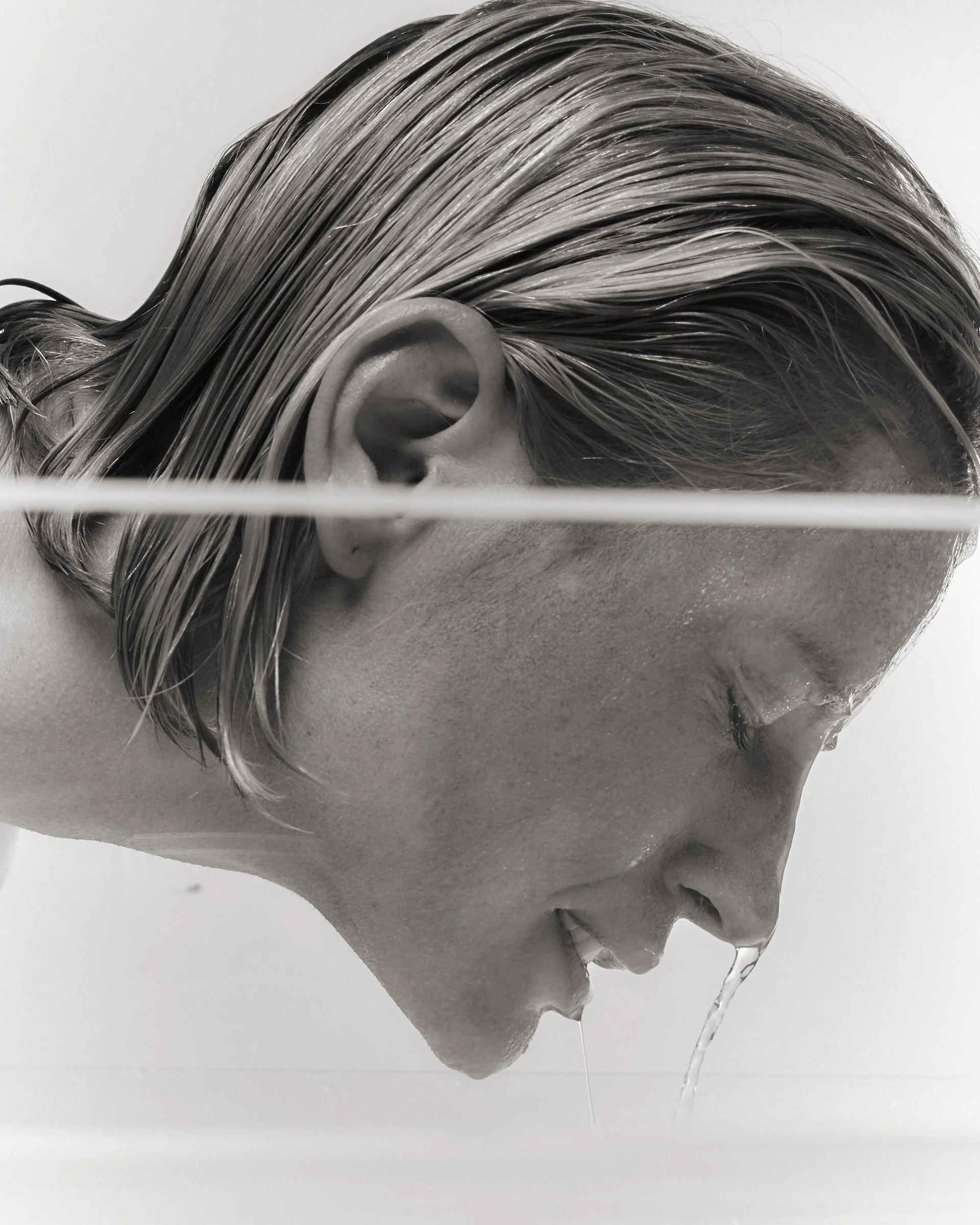 A black and white photograph of a person's head with wet, slicked-back hair, lying on a flat surface, with water streaming from their hair and face.