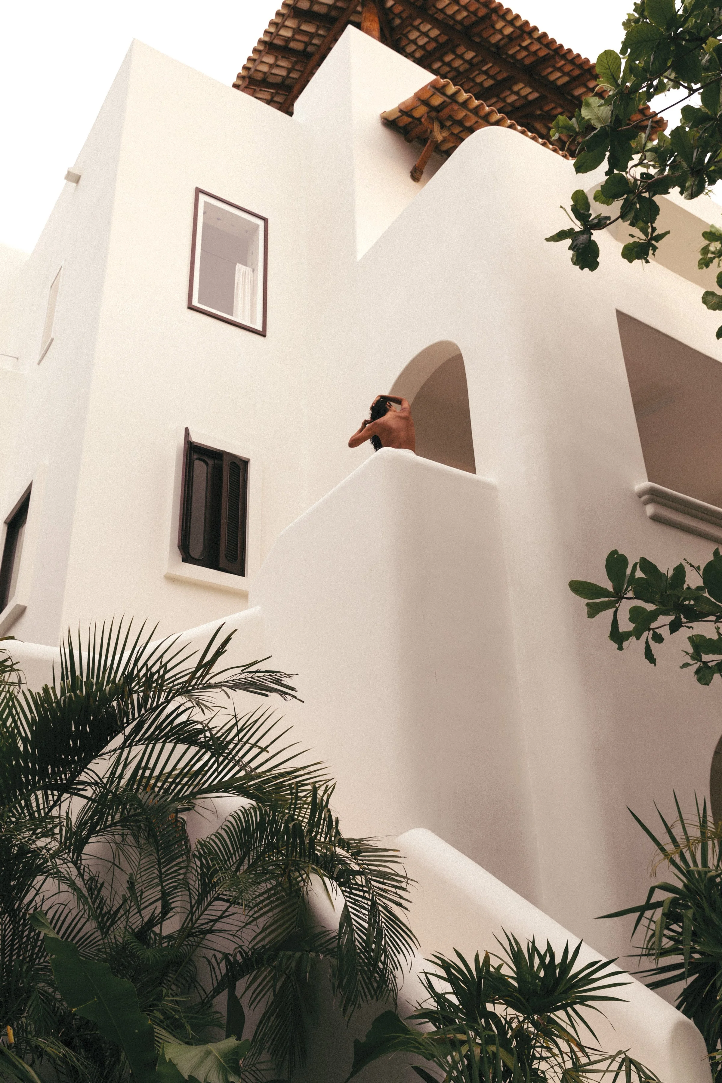A white modern building with small windows, a balcony, and a wooden roof, surrounded by lush green plants and trees.