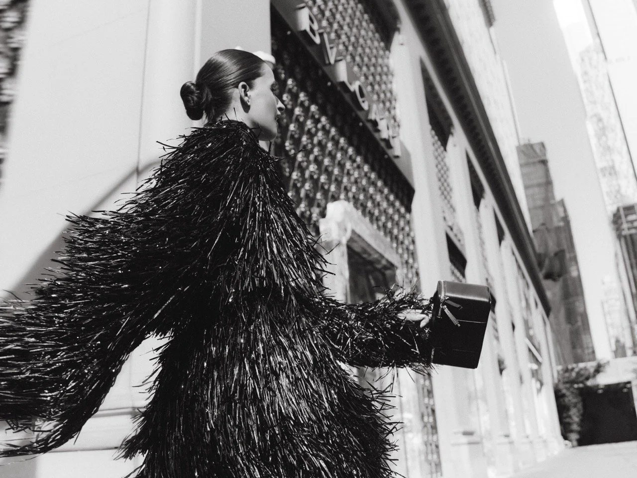 A woman in a shiny black feathered coat walking on a city sidewalk while holding a small handbag.